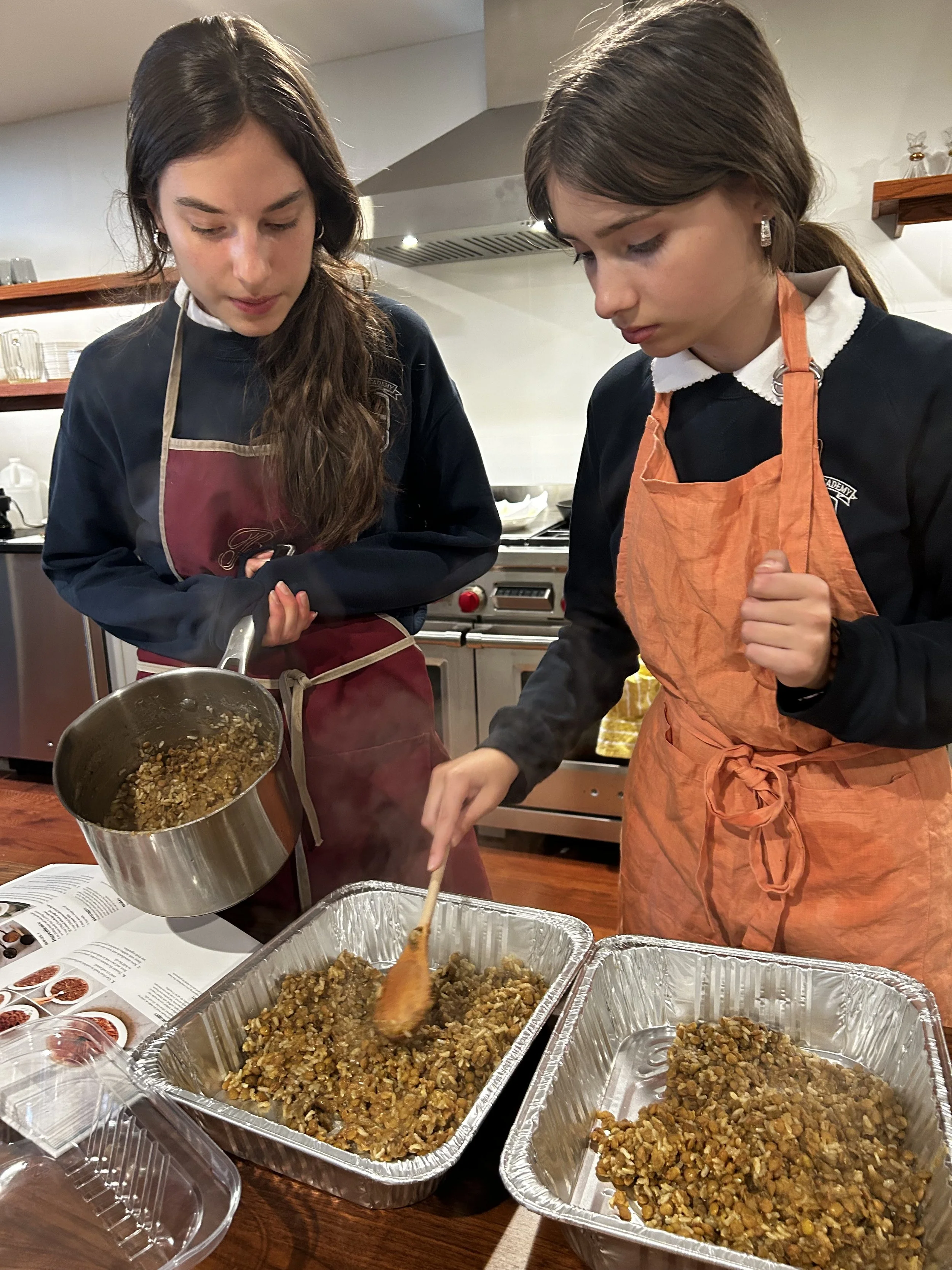 Two young women in an indoor kitchen, wearing aprons, are preparing a large batch of cooked lentils or rice in aluminum trays, with one stirring and the other pouring or adding ingredients from a pot.