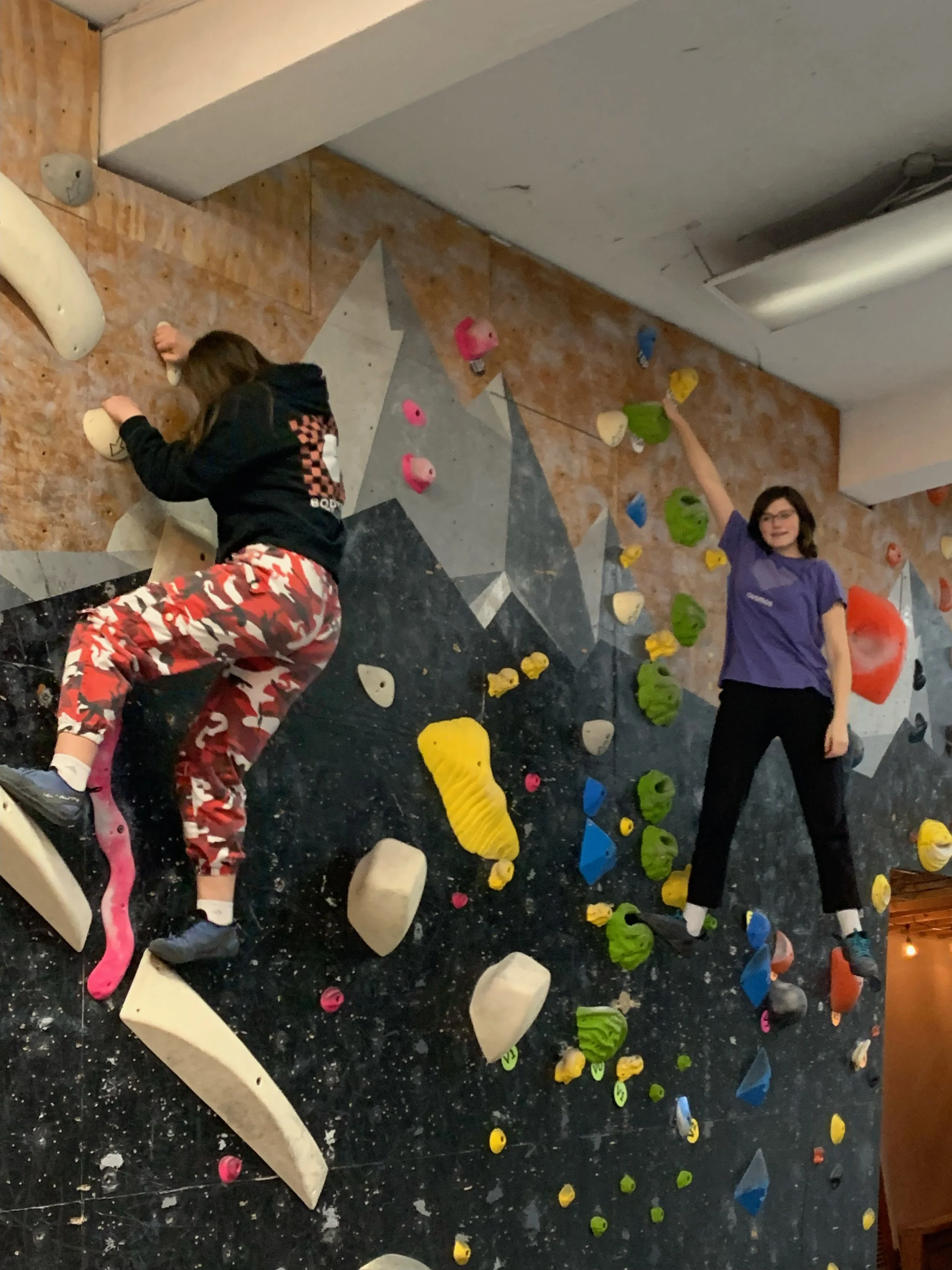 Two girls climbing on an indoor bouldering wall with colorful holds at a gym.