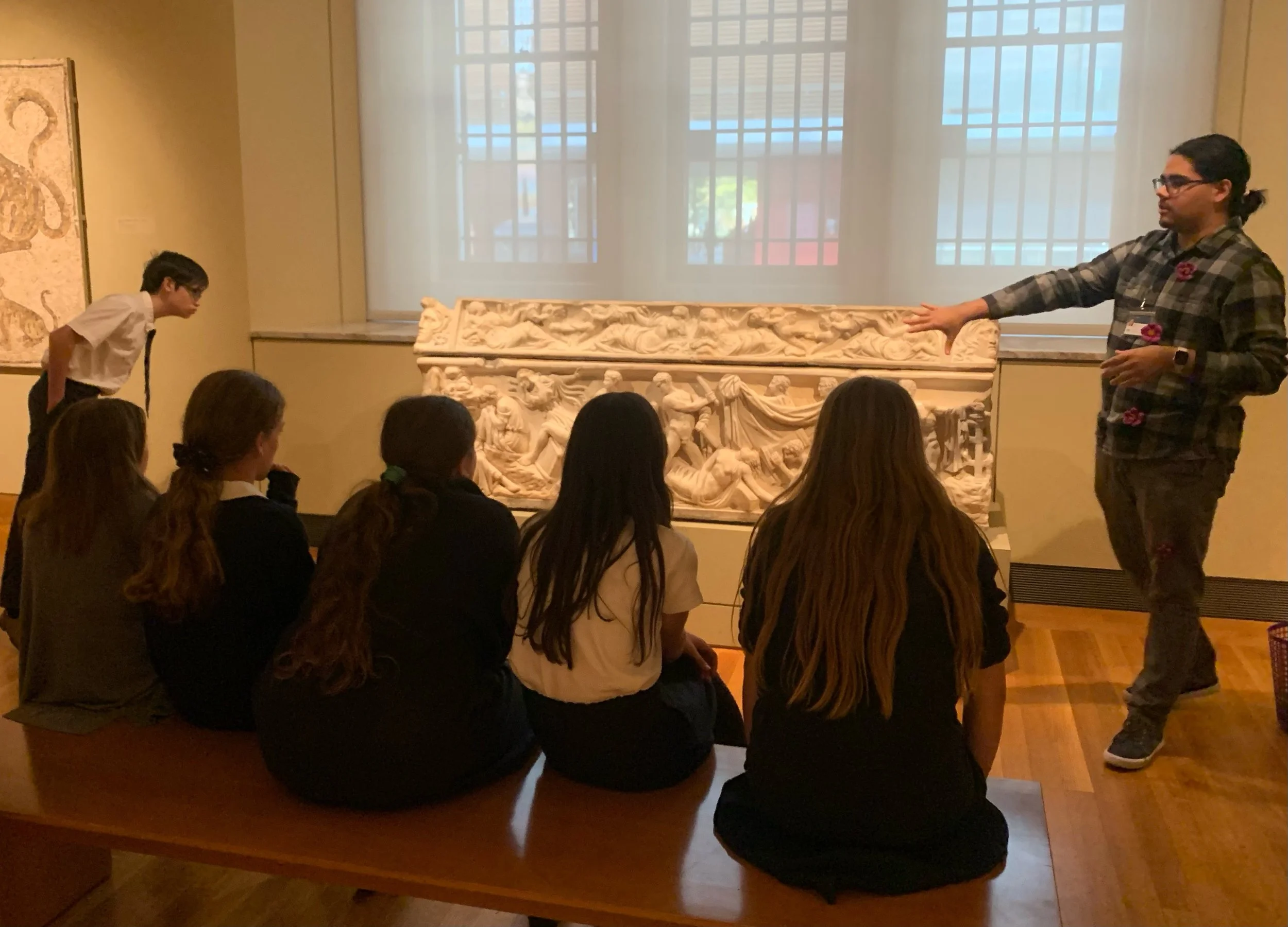 A group of students sitting on a bench in a museum, listening to a guide explaining a marble relief sculpture.