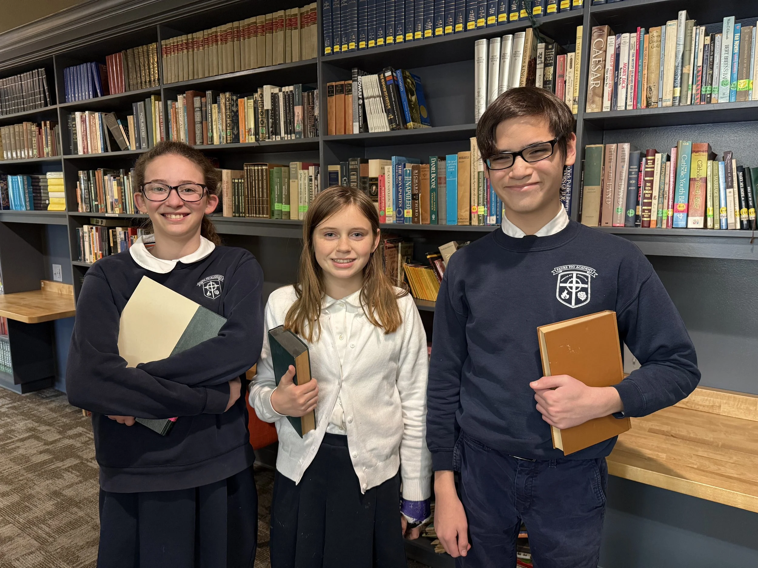Three students, two girls and one boy, stand in front of a bookshelf filled with books in a library. All are holding books and smiling at the camera.
