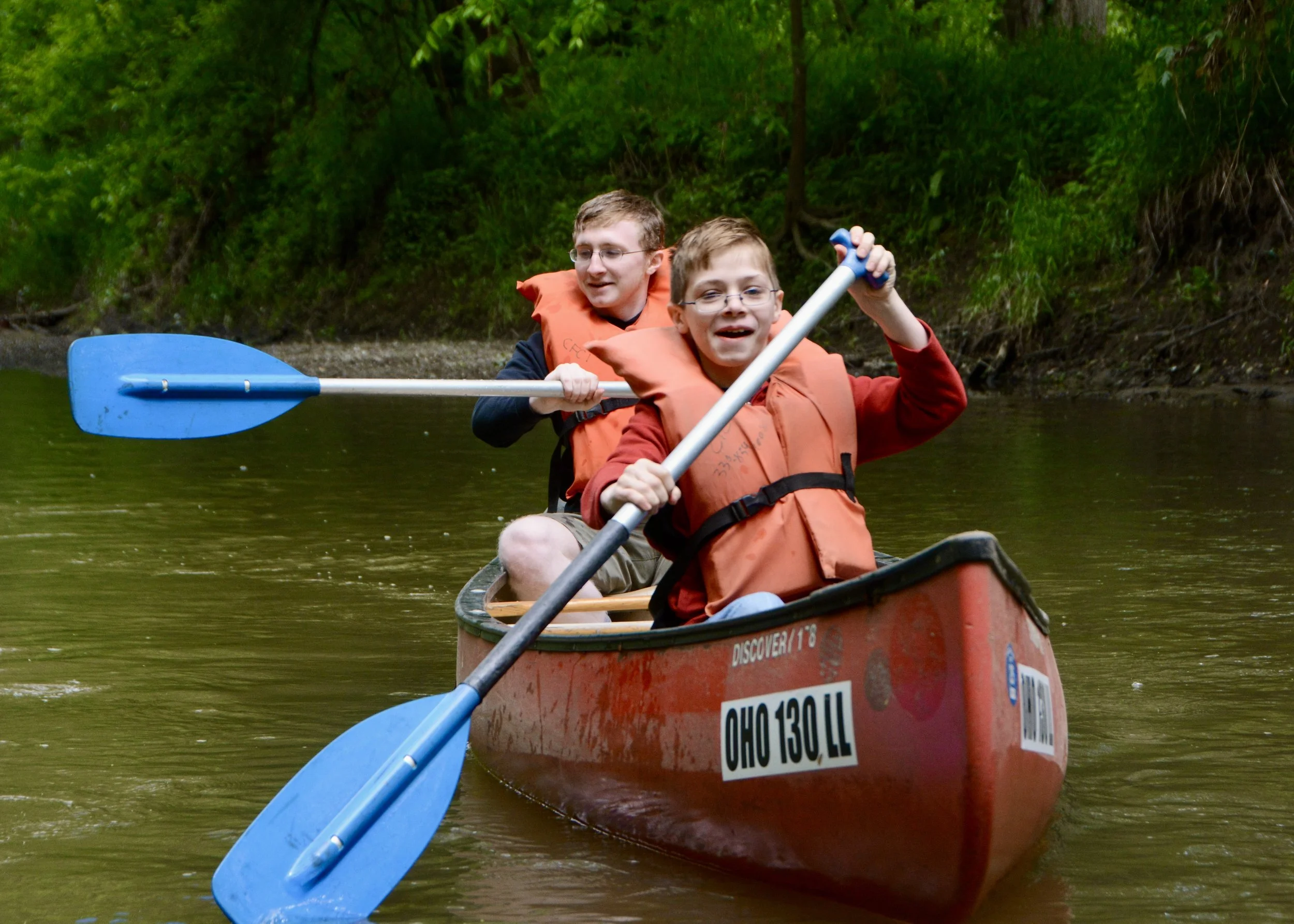 Two boys kayaking on a river surrounded by green trees, wearing orange life jackets and holding paddles