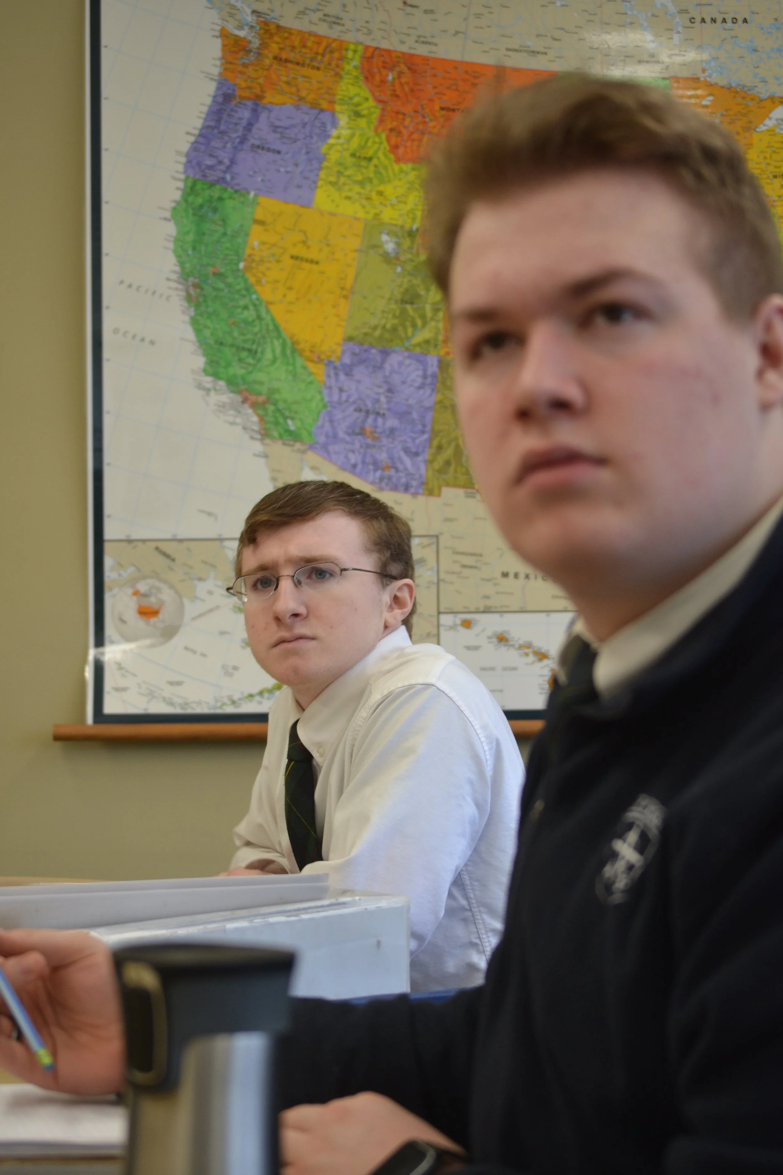 Two young male students sitting in a classroom with a large color-coded map of the United States on the wall behind them. One student wearing glasses and a white shirt is looking slightly to the side, while the other in a black jacket is turned more toward the camera.