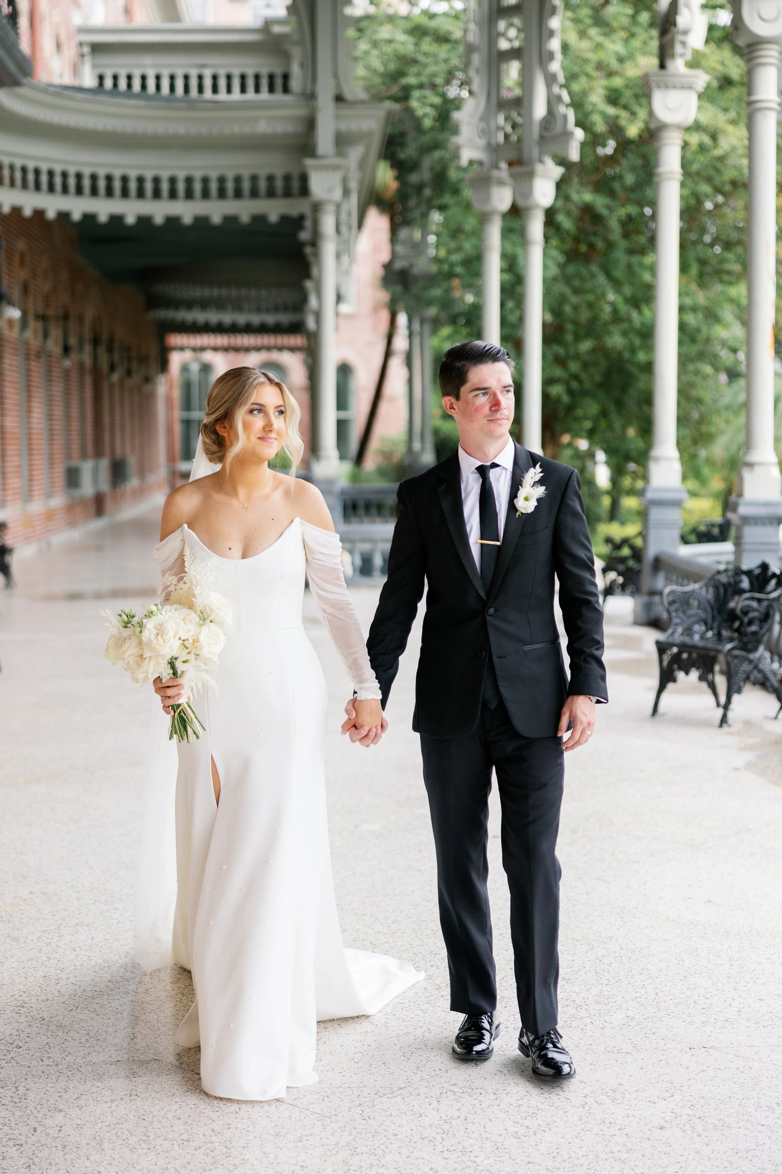 A bride and groom walking hand in hand outdoors on a covered porch at the University of Tampa
