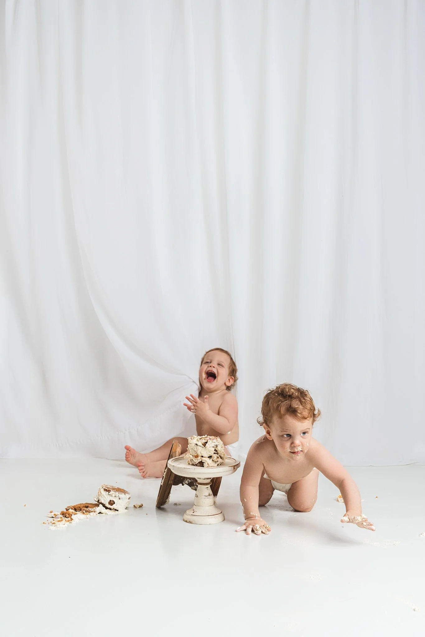 First birthday cake smash moment with two babies reacting differently, one laughing while the other crawls away during a relaxed, baby-led milestone session