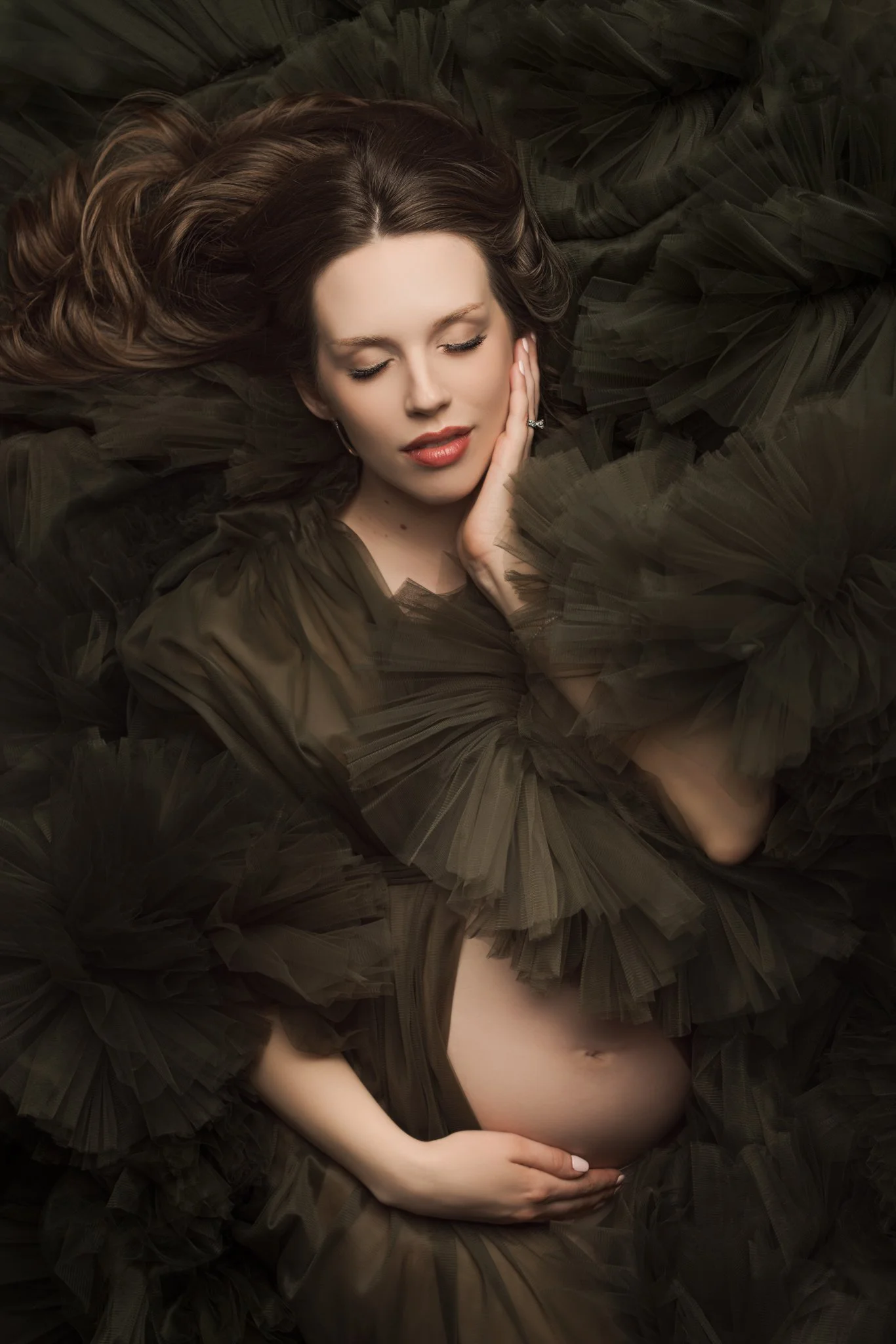 Fine art maternity portrait of an expecting mother wearing a flowing tulle gown, gently cradling her baby bump in a dark, moody studio setting, capturing a serene and timeless moment.