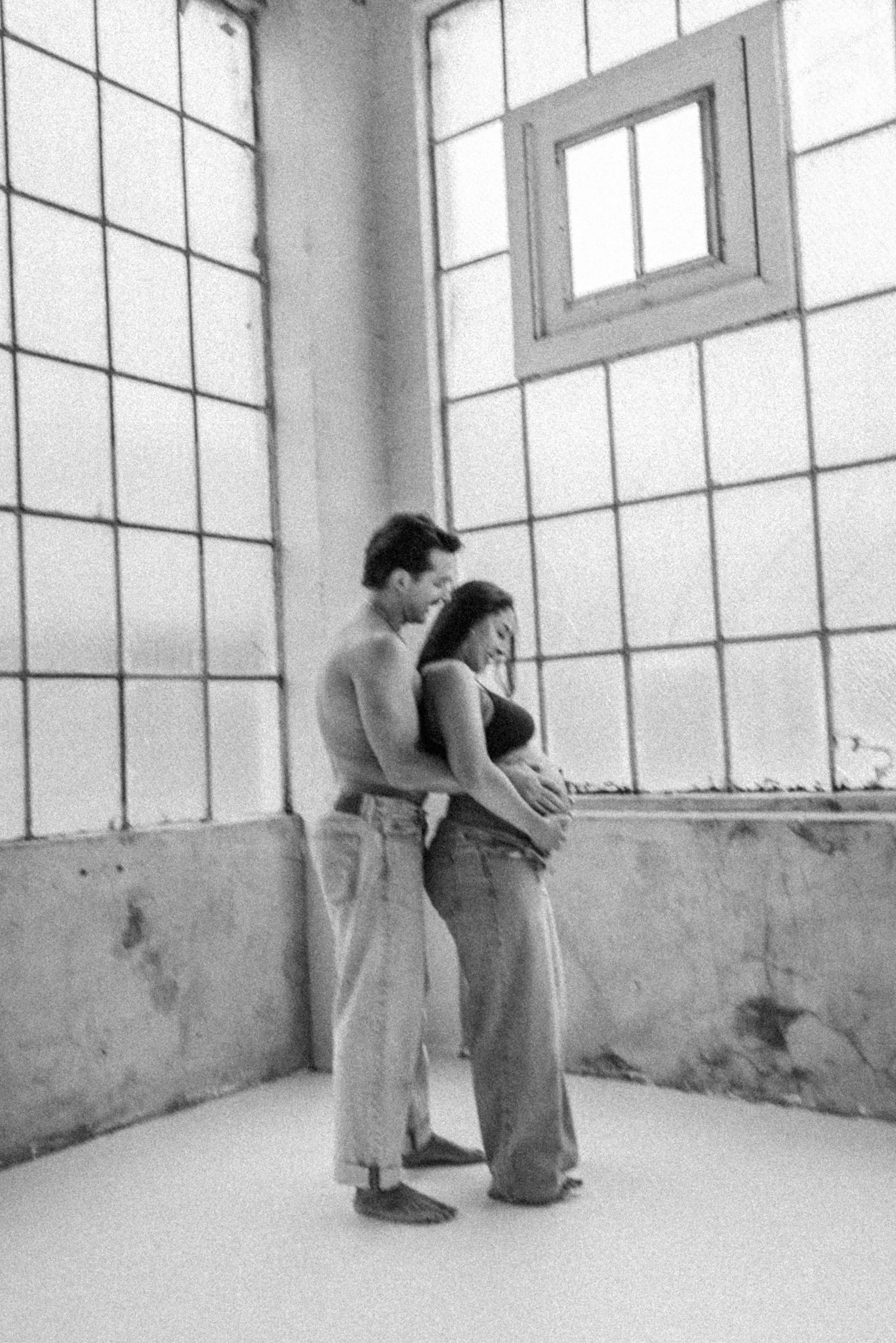 Black and white maternity portrait of an expecting couple standing close together in a bright studio, with the partner gently holding the mother’s baby bump, capturing an intimate and timeless moment.