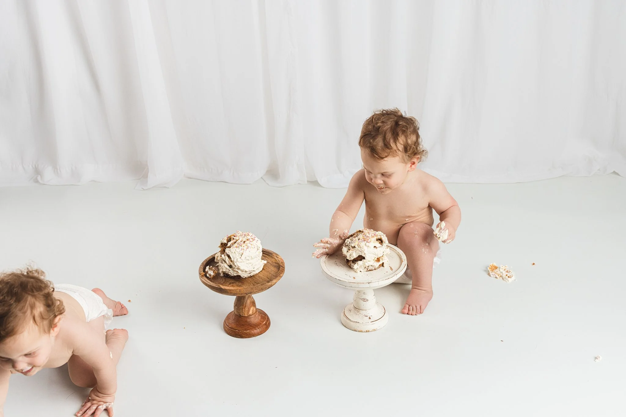 First birthday cake smash moment with two babies reacting differently, one laughing while the other crawls away during a relaxed, baby-led milestone session