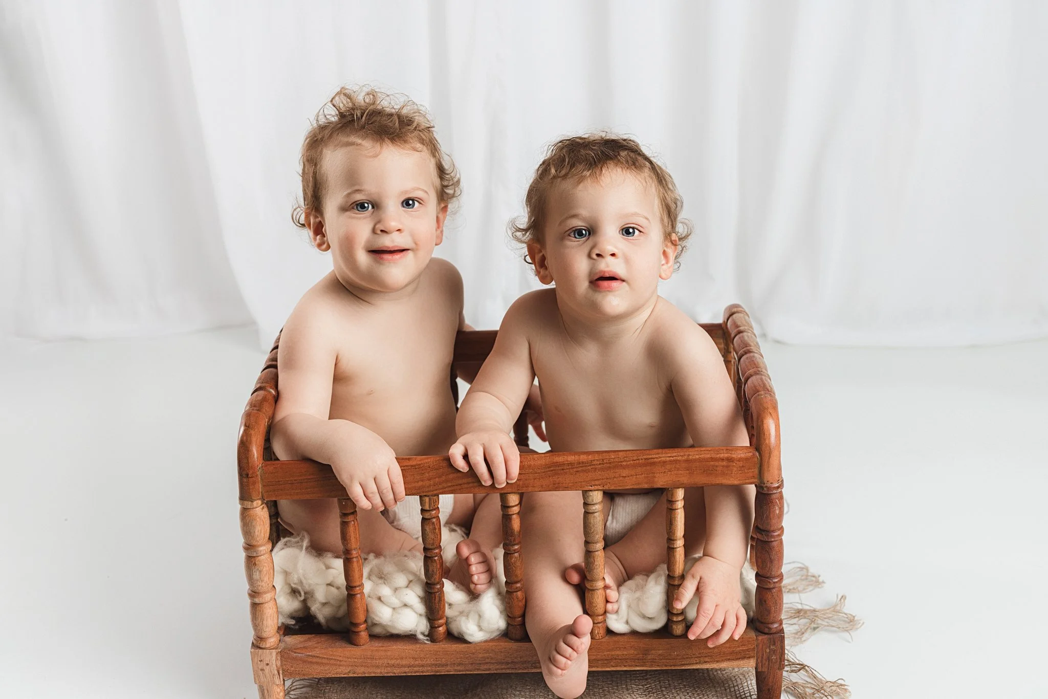 Twin babies sitting together in a wooden bench during a milestone photo session, captured in a bright, minimal studio setting, showcasing their sweet expressions and sibling bond.