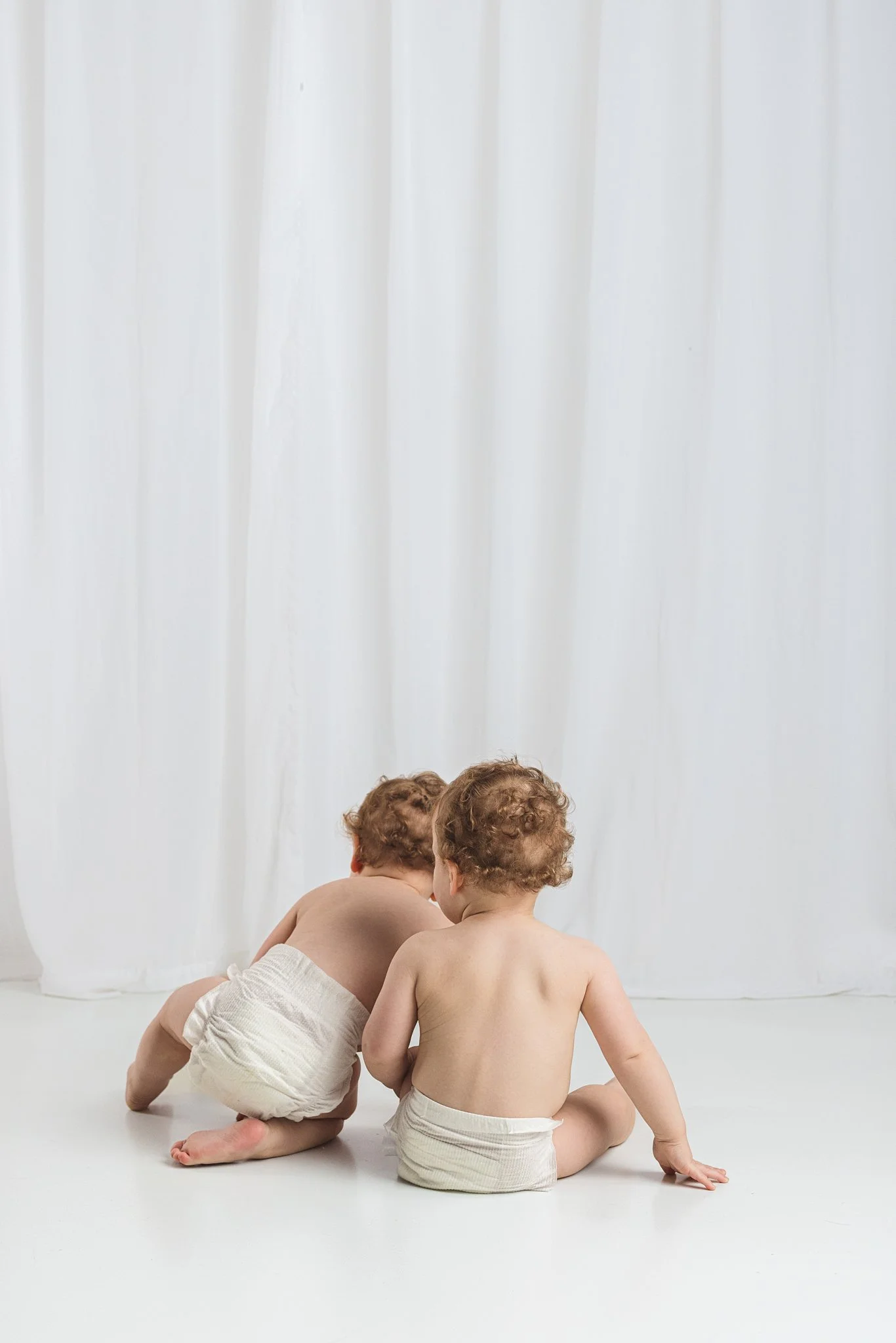 Two babies sitting side by side with their backs to the camera during a baby-led milestone session, capturing a quiet moment where movement and exploration are always welcome