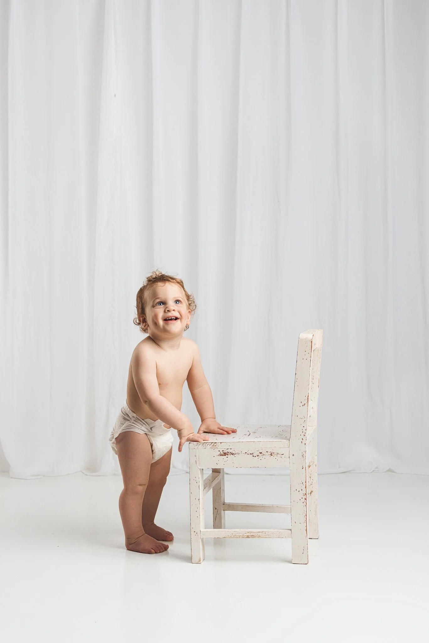 Happy baby standing and holding a small white chair during a first birthday milestone photography session in Lake Tapps