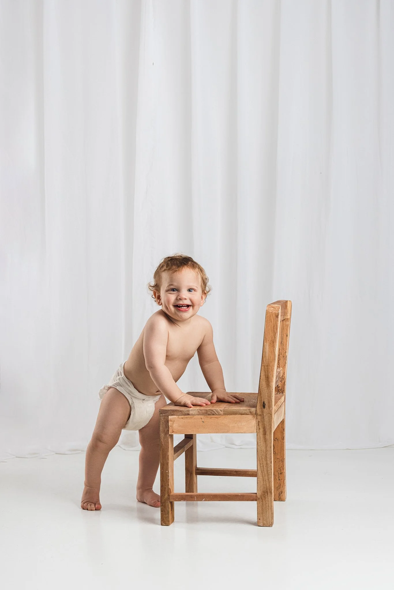 Baby standing and smiling while holding onto a wooden chair during a first birthday milestone session in Lake Tapps, Washington