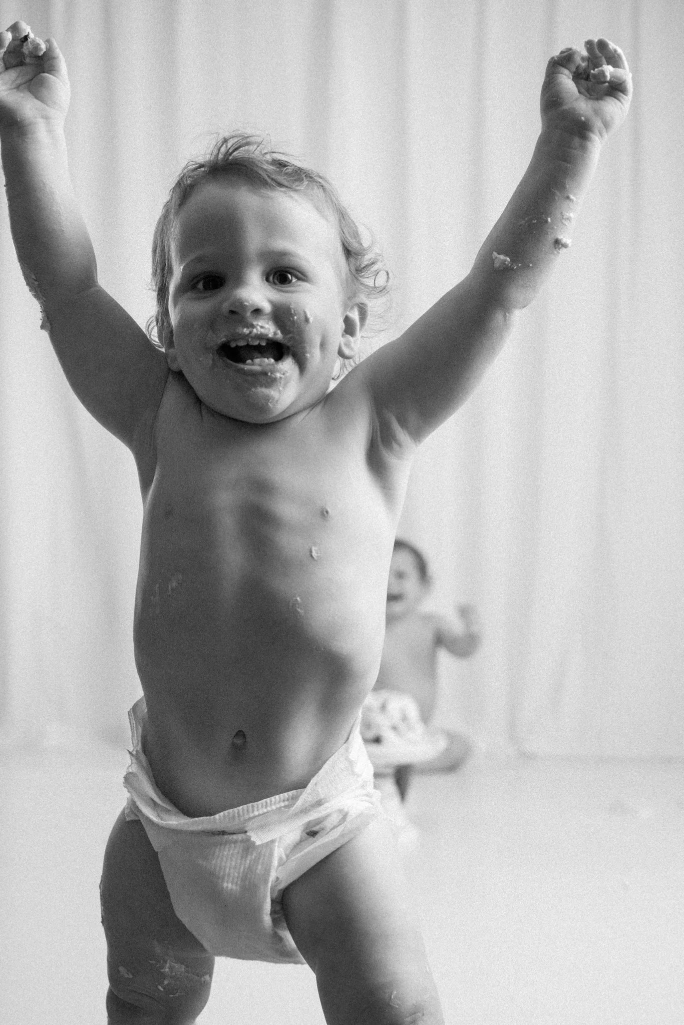 Black and white photo of a baby with arms raised and frosting on their face during a first birthday cake smash session in a studio