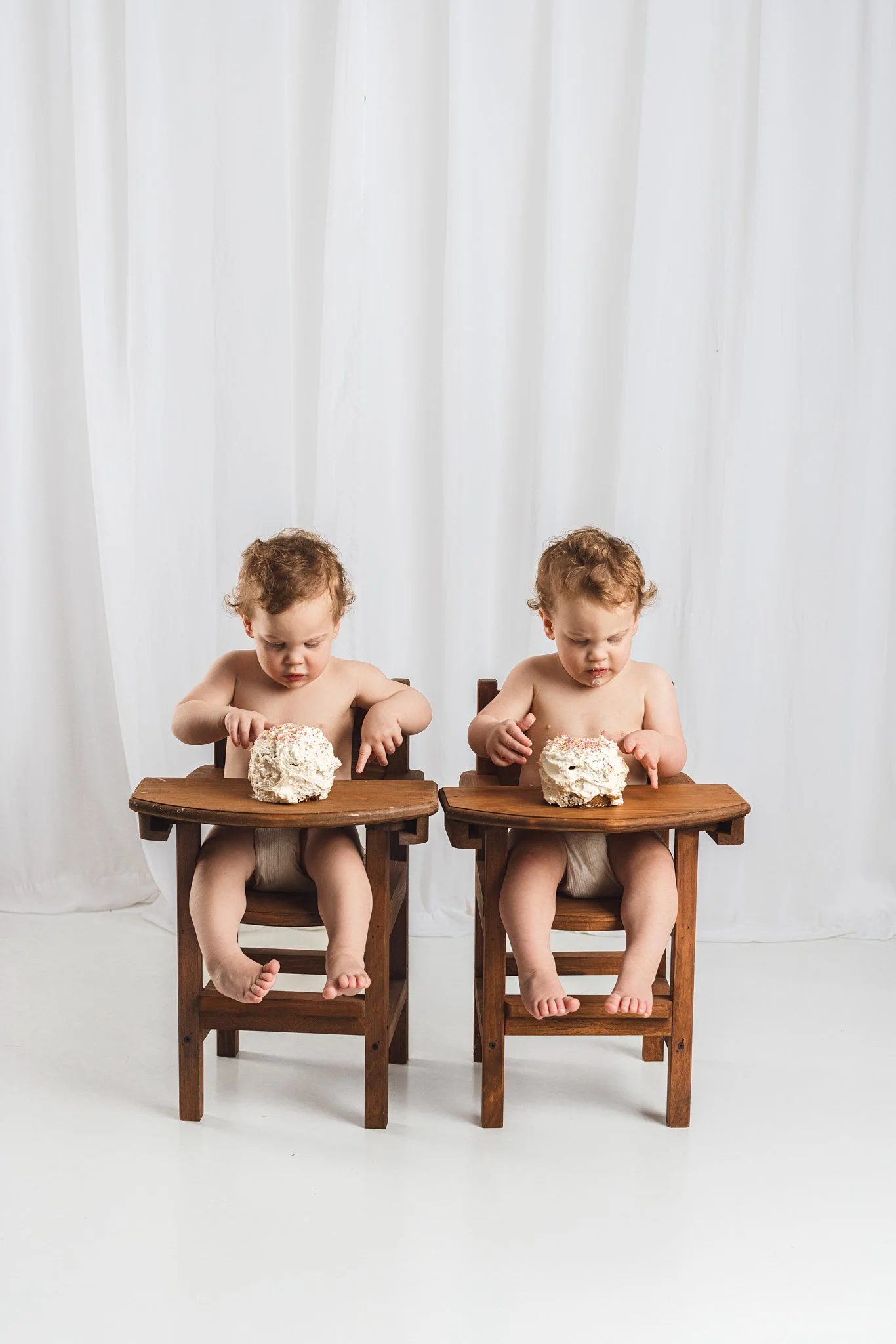 Twin babies seated in wooden high chairs exploring their first birthday cakes during a studio cake smash session in Seattle