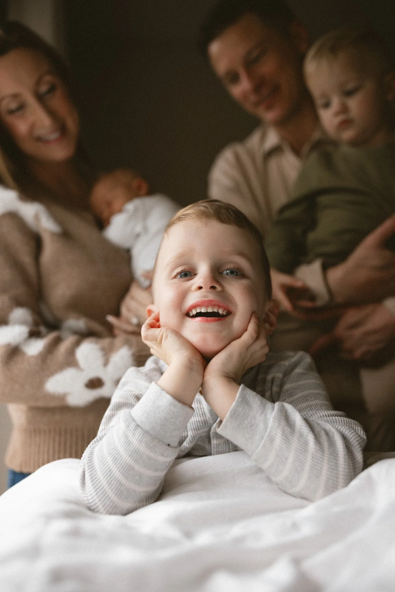 A smiling toddler poses on a bed during a cozy in-home newborn family session, with parents holding a newborn and another child in a softly lit bedroom.