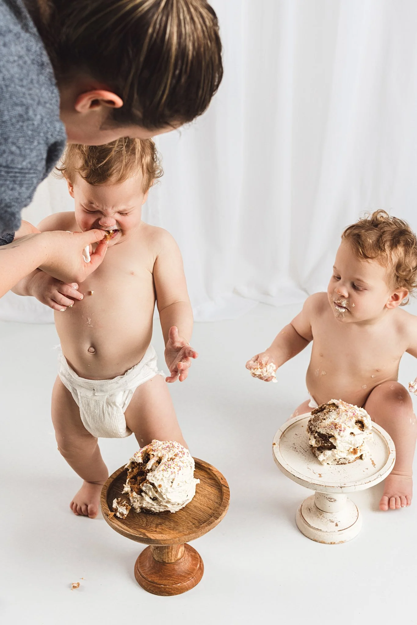 Parent helping baby taste cake during a first birthday cake smash session while another baby sits nearby in the studio