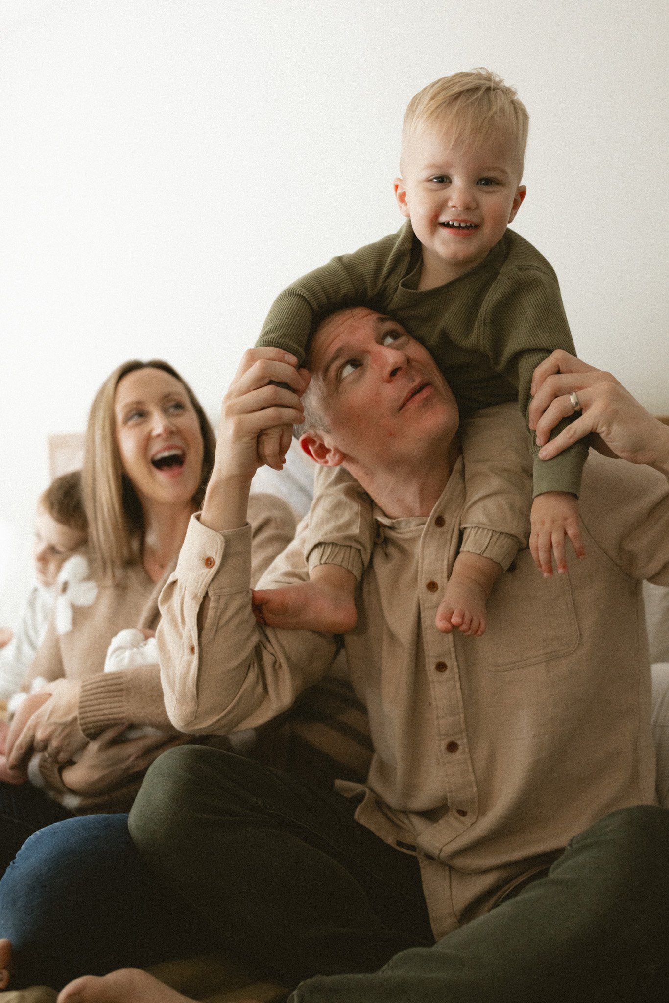 A father lifts his smiling toddler onto his shoulders while a mother holds their newborn during a cozy in-home lifestyle family photography session in West Seattle.