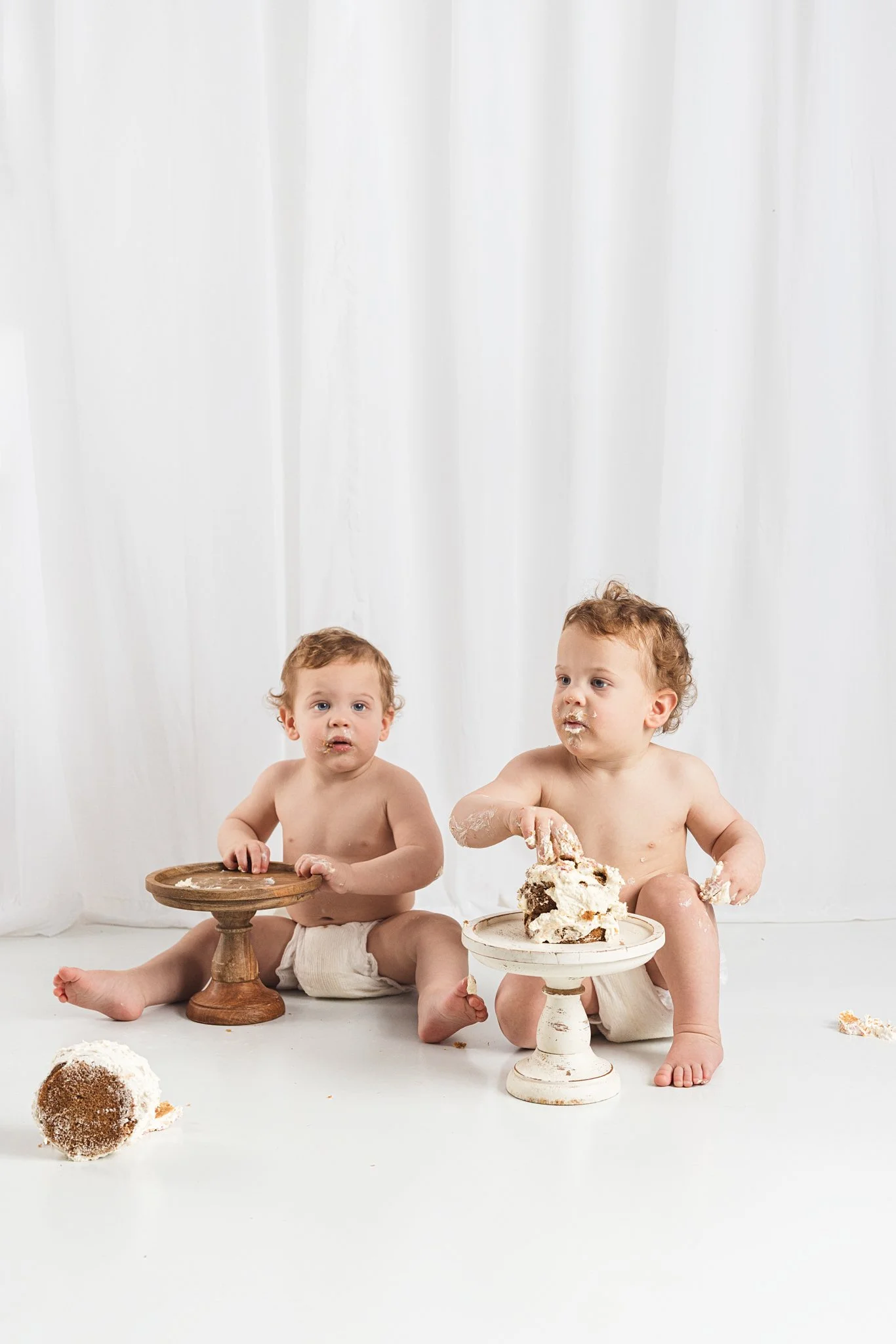 Two babies sitting side by side during a first birthday cake smash session, exploring their cakes at their own pace with frosting-covered hands in a relaxed studio setting
