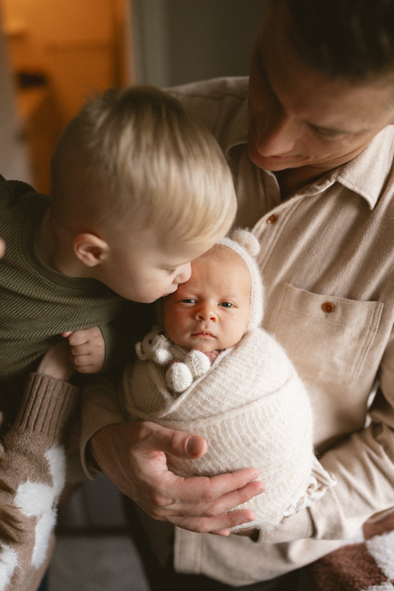 A toddler gently kisses his newborn sibling while their father holds the baby during a cozy in-home newborn photography session in Seattle.