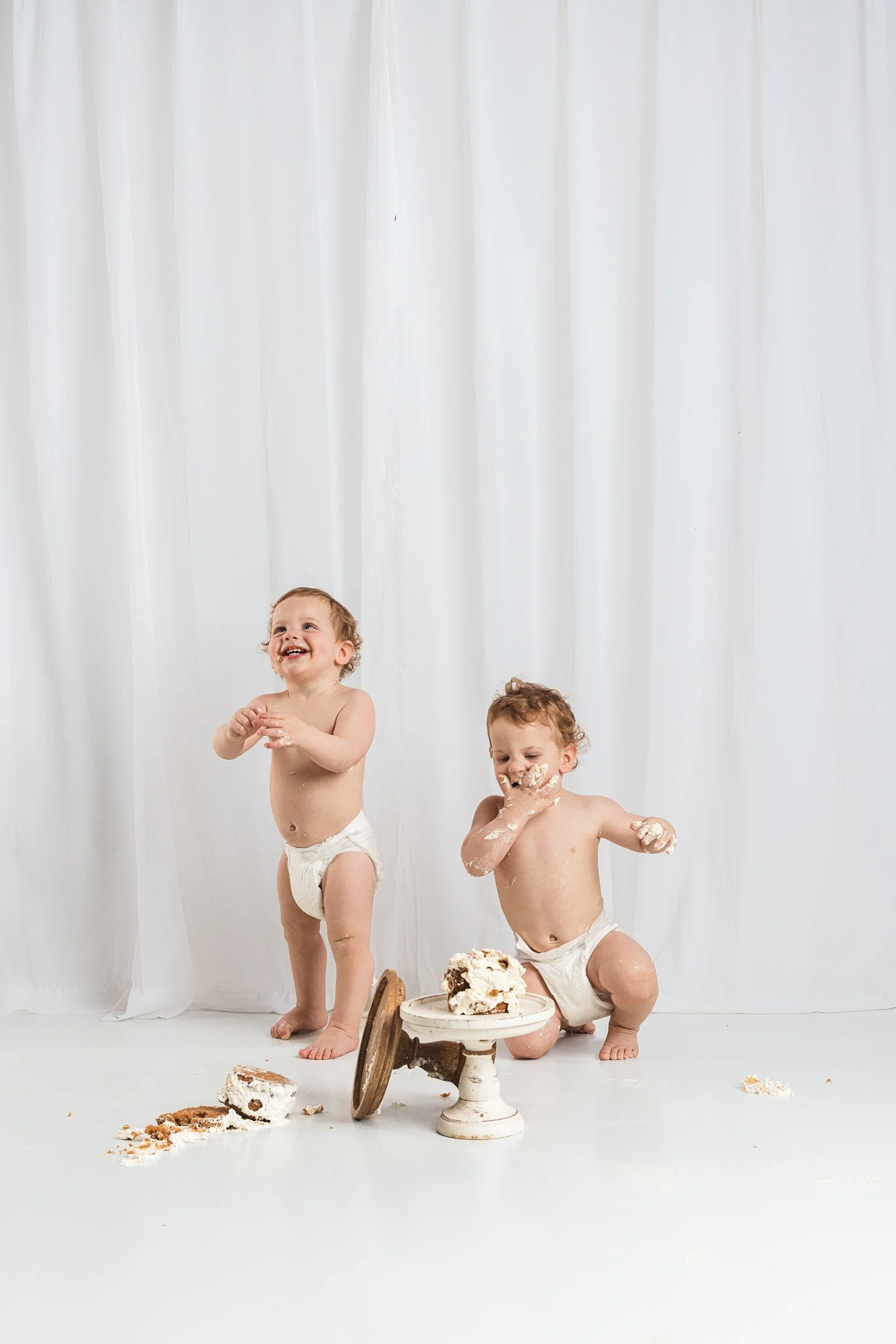 Two babies standing during a first birthday cake smash session, exploring cake in their own way with frosting-covered hands in a baby-led studio environment