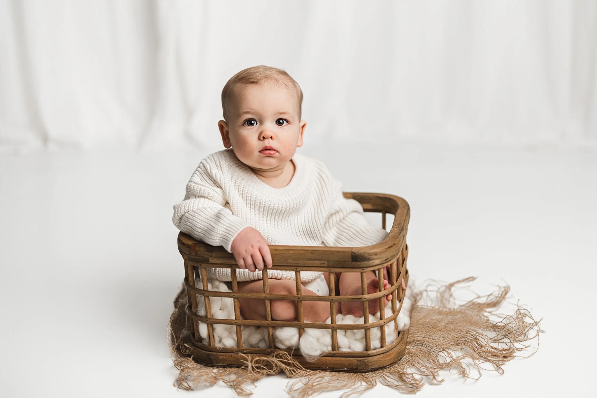 Baby sitting in a wooden basket during a studio milestone photography session with a soft, neutral backdrop.