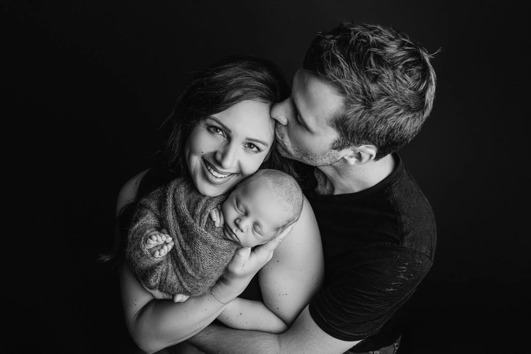 Black and white portrait of a smiling mother holding her newborn baby wrapped in a cozy blanket, while the father gently kisses the mother’s temple, capturing a tender family moment in a studio setting