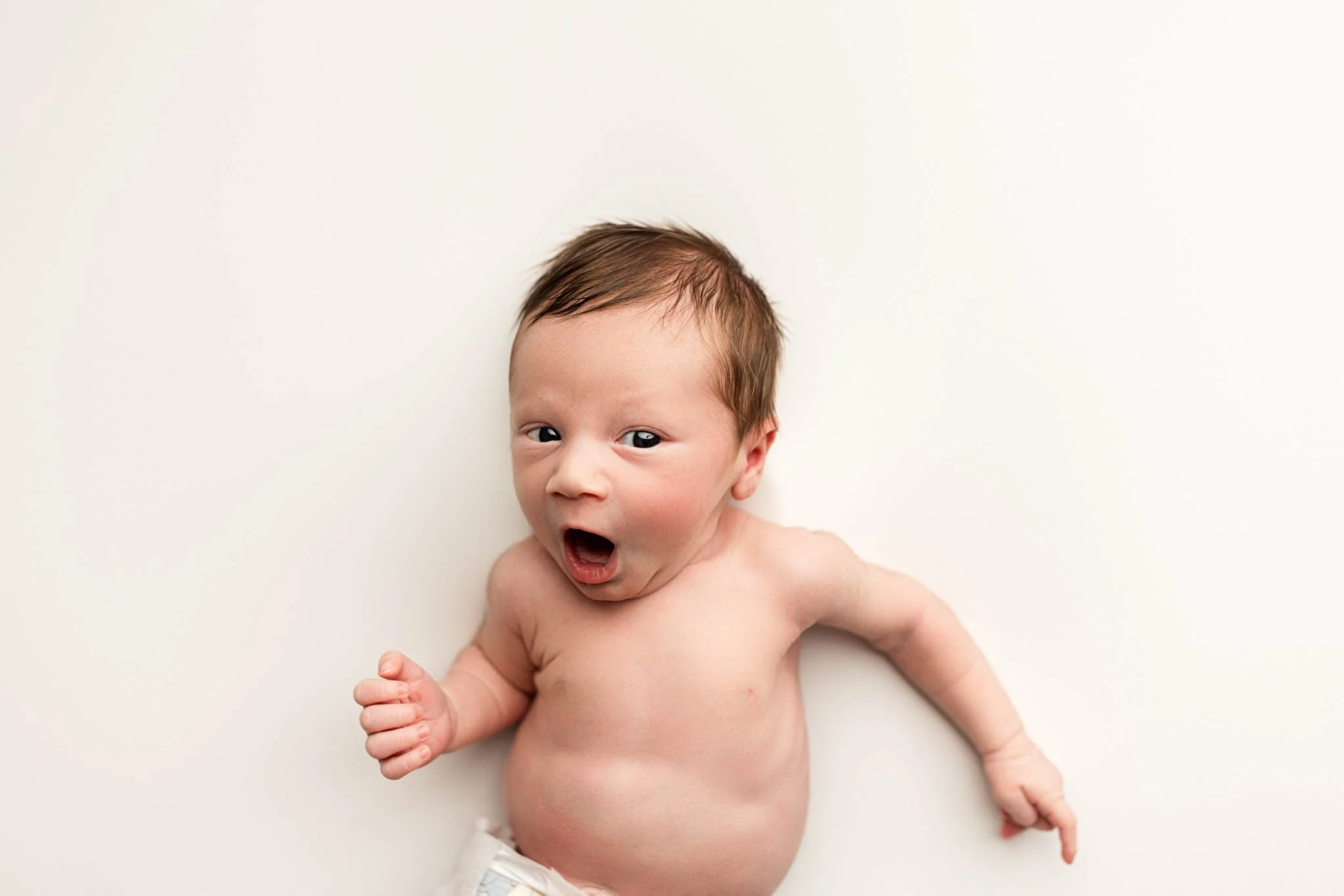 Newborn baby with dark hair yawning while laying on a soft white background during a professional studio newborn photography session in Seattle.