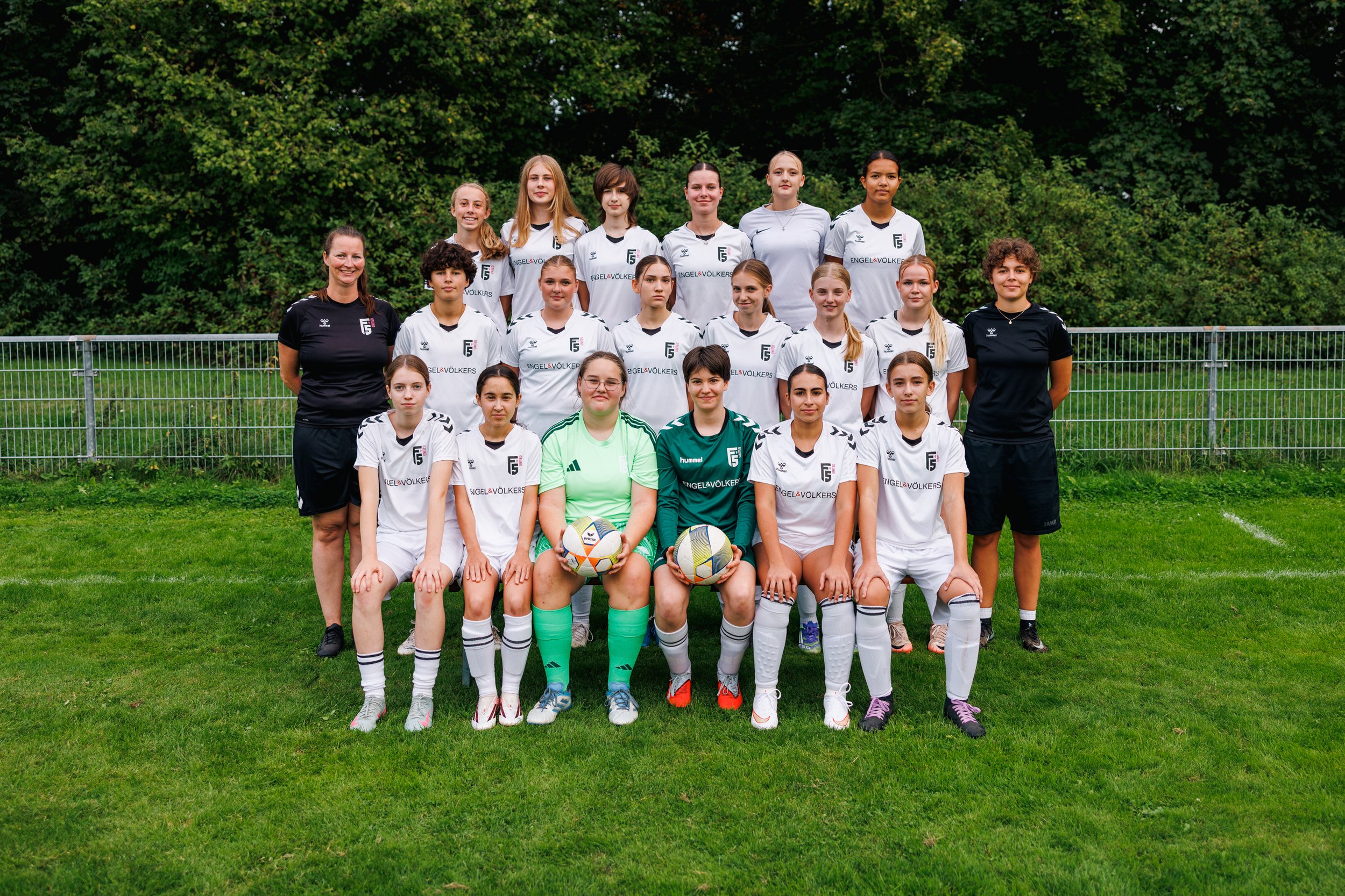 Mädchenfußballteam mit Trainerinnen posiert auf dem Spielfeld, alle tragen weiße Trikots mit schwarzen Details, zwei Mädchen in Torwartkleidung halten Bälle, im Hintergrund dunkle grüne Bäume.