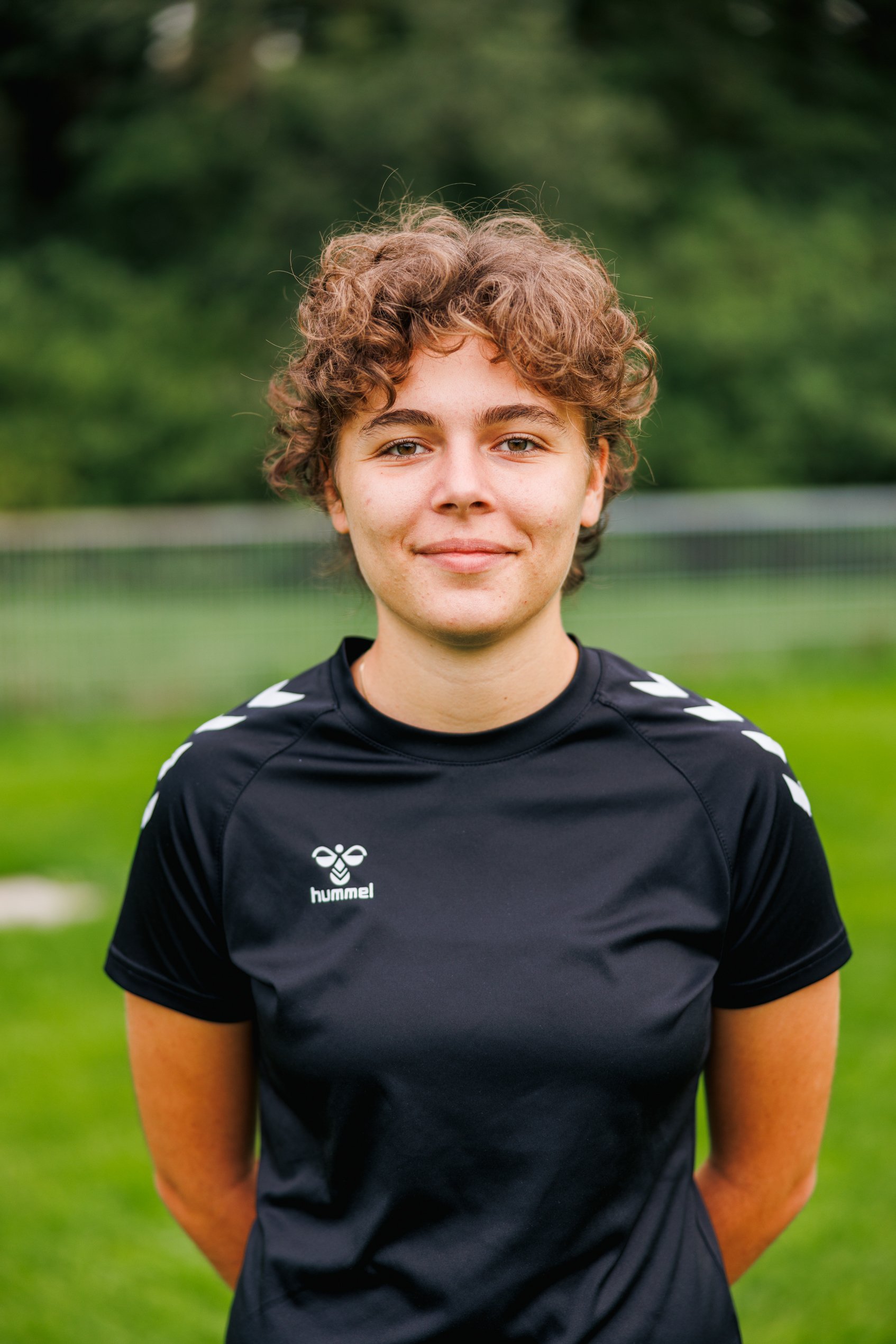 Porträt einer jungen Frau mit lockigem braunem Haar, der auf einem Fußballfeld steht, trägt ein schwarzes Sporttrikot von Hummel, im Hintergrund grüne Bäume und eine Zaunlinie.