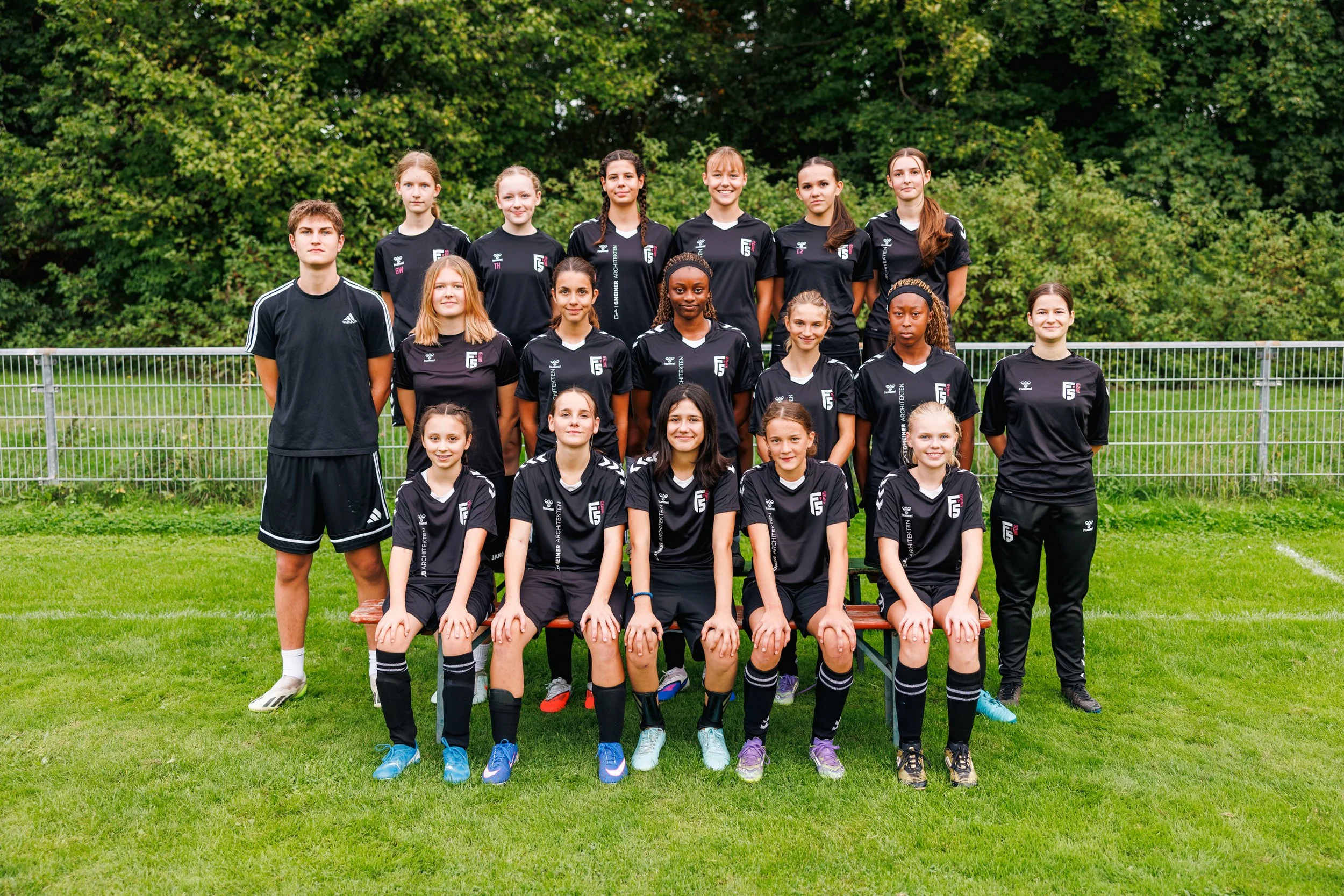 Gruppenfoto von jungen Frauen und einem jungen Mann in schwarzen Fußballtrikots auf einem Rasenfeld mit Bäumen im Hintergrund.