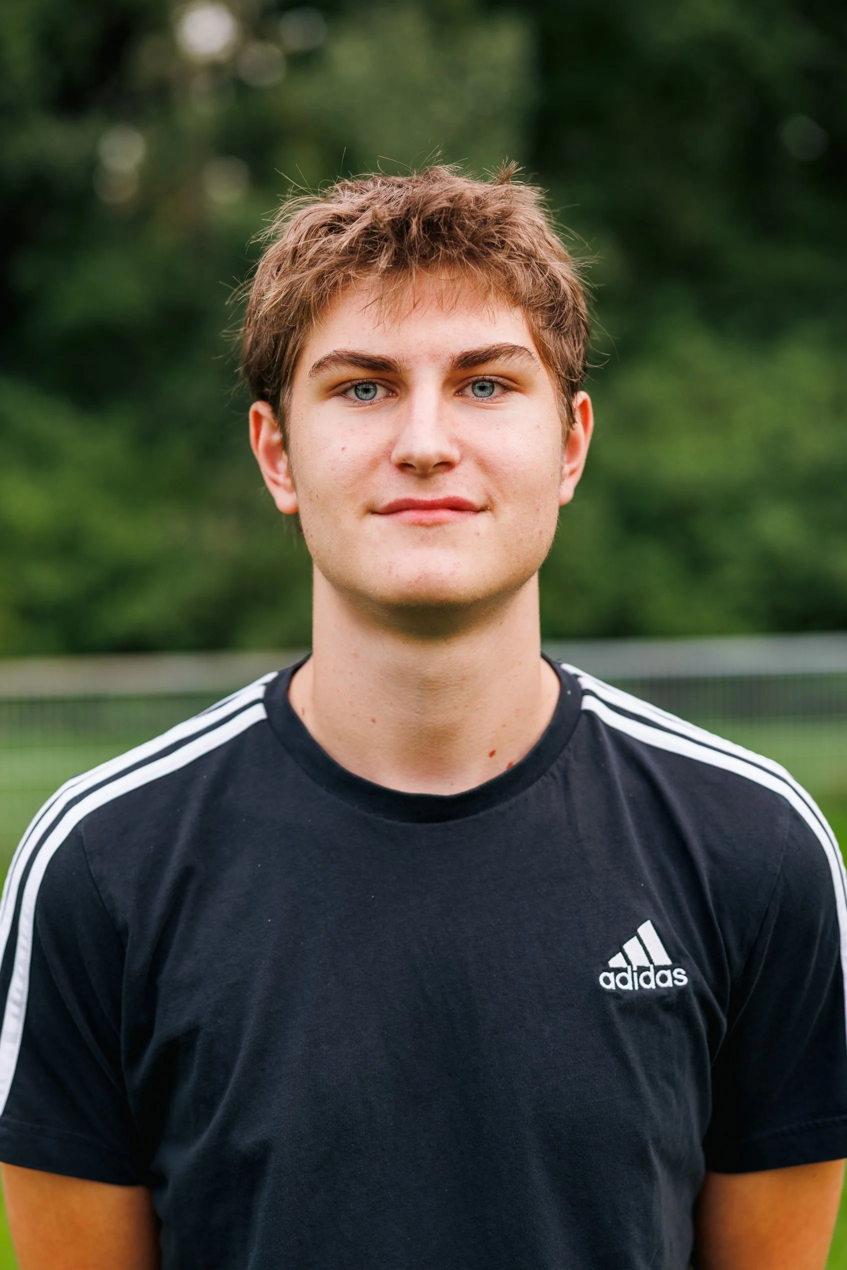 Junge mit blauen Augen, braunem Haar und schwarzem Adidas-T-Shirt auf einem Fußballfeld im Freien.