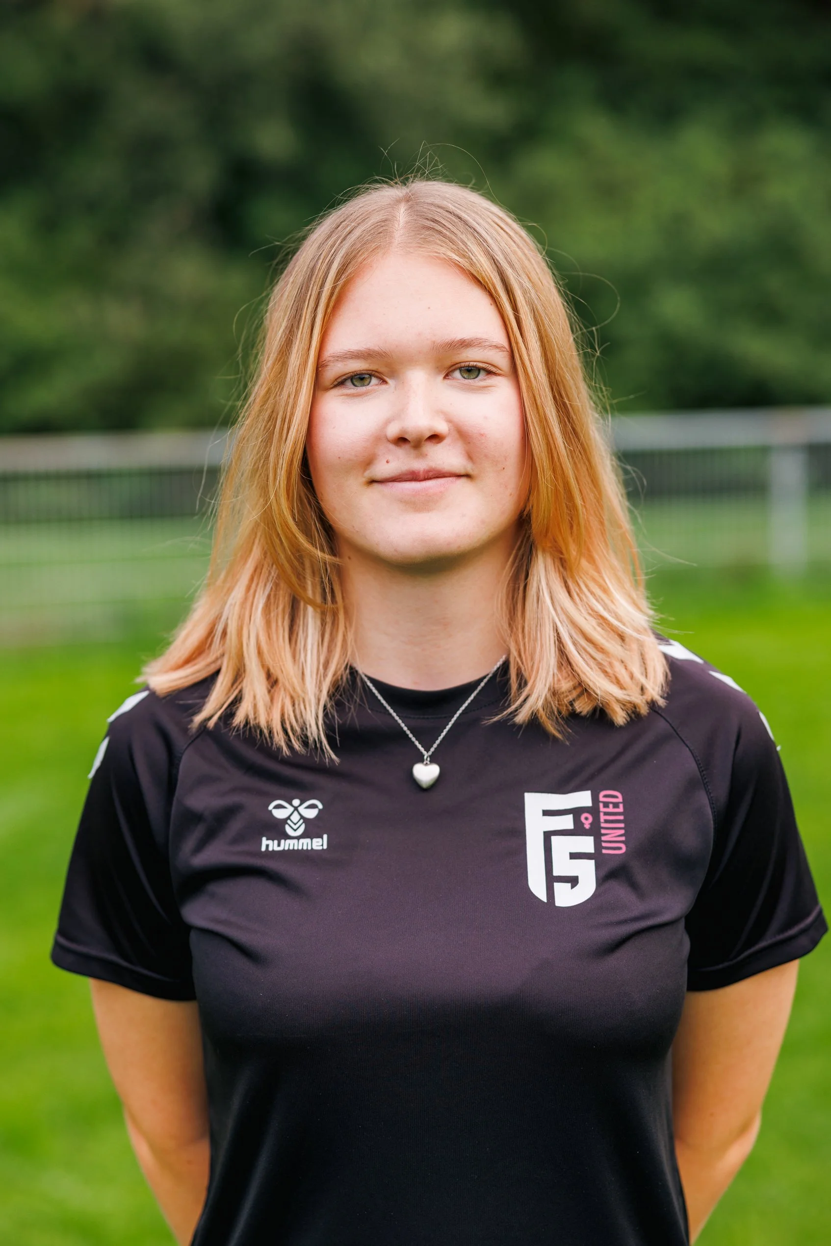 Junge Frau mit roten Haaren in einem schwarzen Sporttrikot, stehend im Freien auf einem Fußballfeld mit grünem Hintergrund, lächelt leicht.