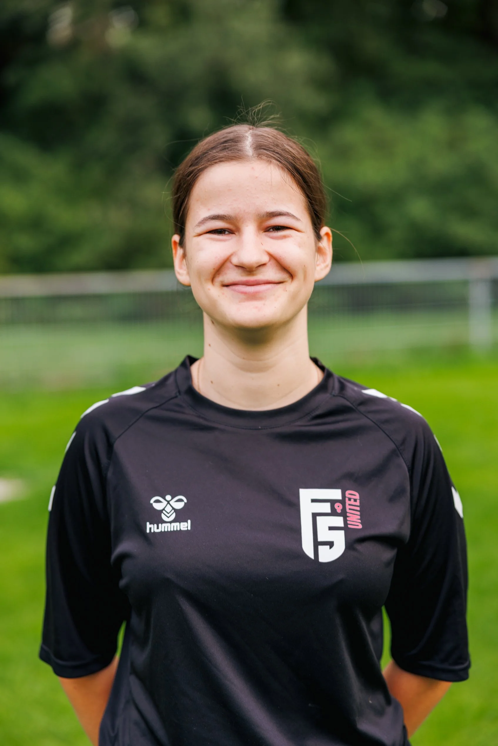 Nahaufnahme eines lächelnden jungen Mädchen in einem schwarzen Sporttrikot auf einem Fußballfeld mit grünem Hintergrund.