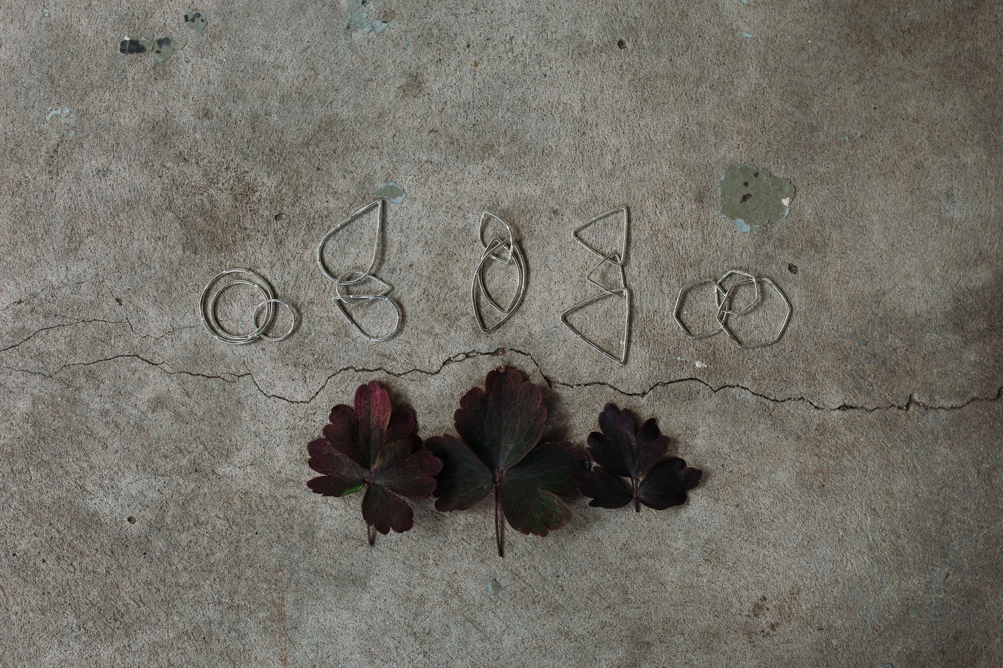 The primary line; Sphere, Drop, Leaf, Mountain and Crystal. (ID: Five silver pendants lie on a cracked concrete floor, just above three dark red aquilegia leaves)