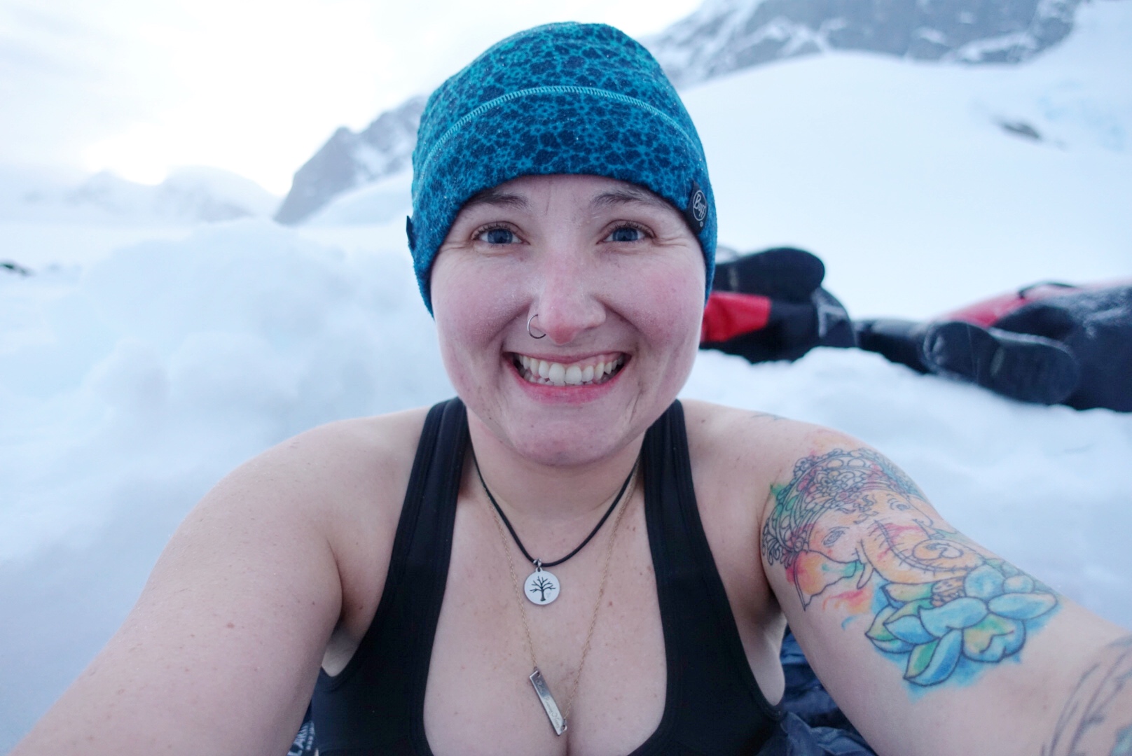 Camping in Antarctica: a woman surrounded by snow smiling at the camera with a blue hat and black sports bra on.