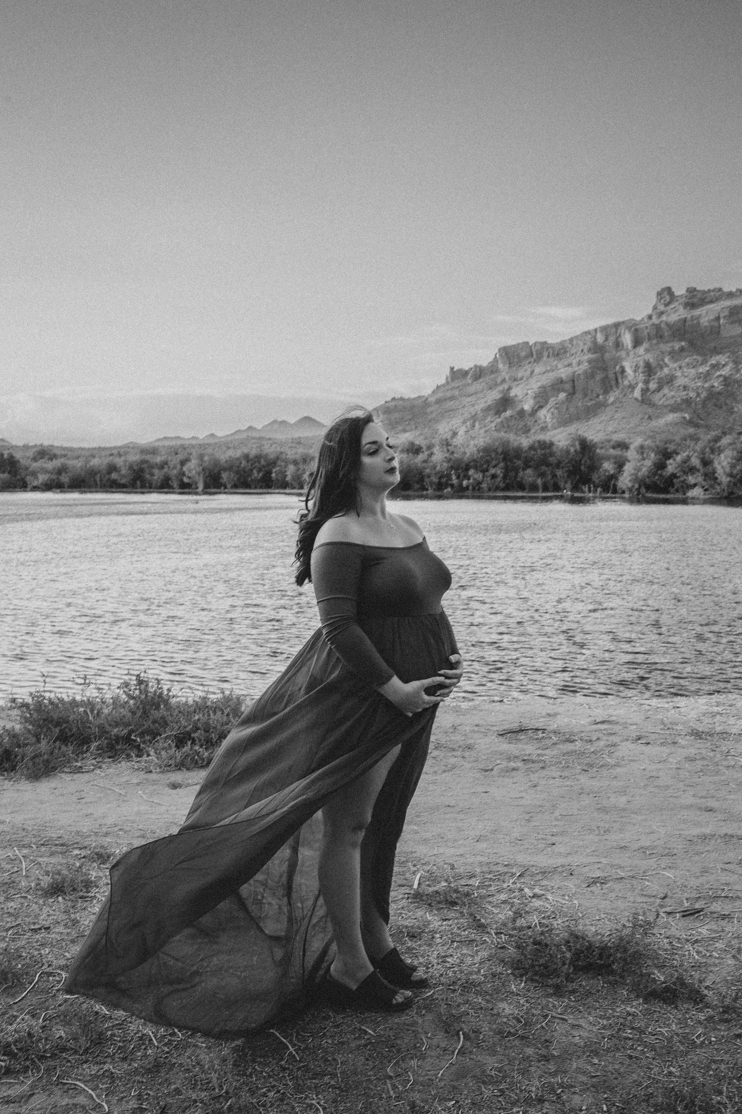 Pregnant woman stands in a dress posing for maternity photos in front of the Salt River and mountain by the best Phoenix maternity photographer in Arizona; Jennifer Lind Schutsky 