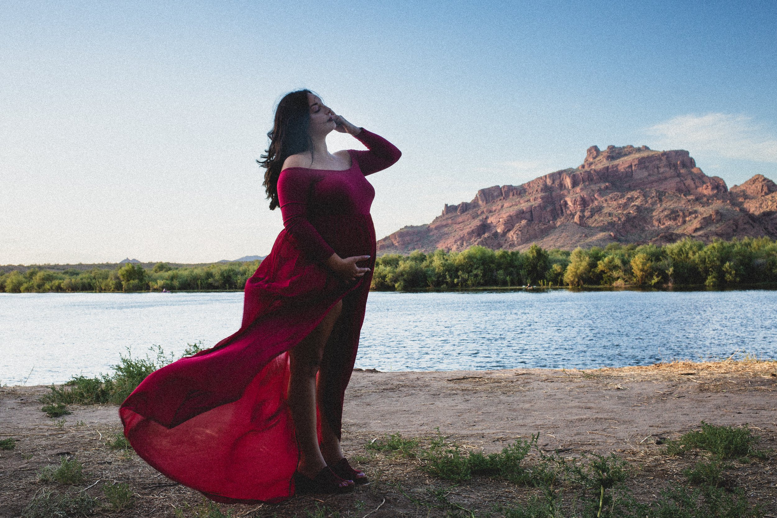 Pregnant woman in a red dress poses for maternity photos in front of the Salt River and mountain by the best maternity photographer in Phoenix; Jennifer Lind Schutsky 