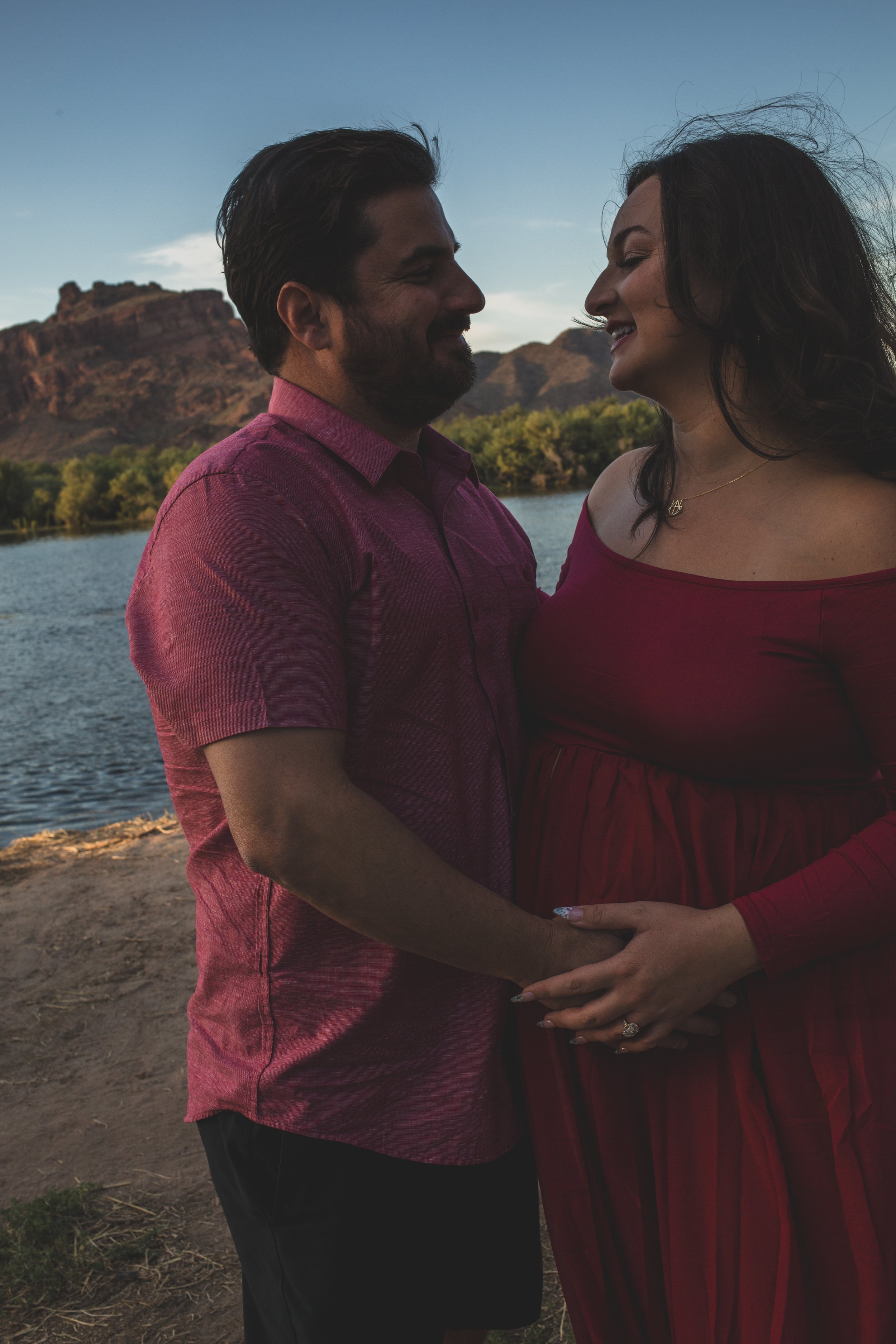 Pregnant woman in a red dress poses with her partner for maternity photos in front of the Salt River and mountain by the best maternity photographer in Phoenix; Jennifer Lind Schutsky.