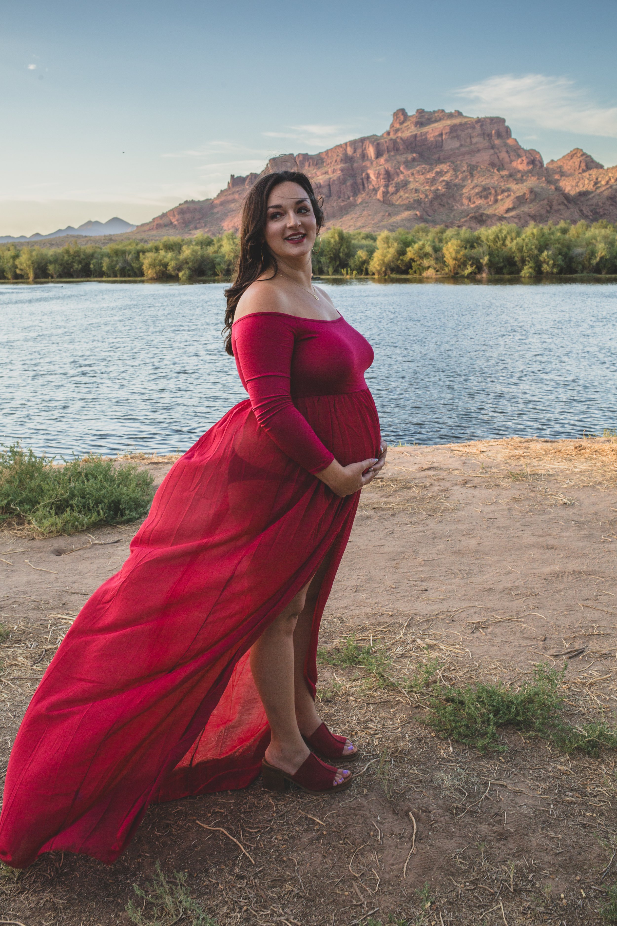Pregnant woman stands in a red dress for candid maternity photos in front of the Salt River and mountain by the best maternity photographer in Phoenix; Jennifer Lind Schutsky.