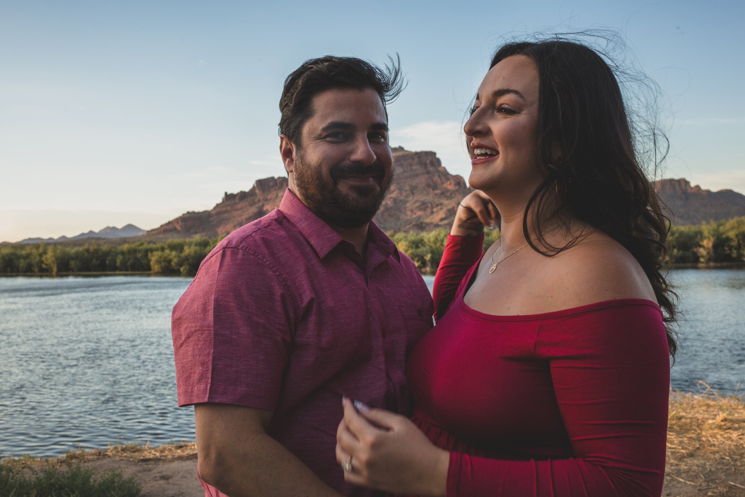 Pregnant woman stands with her partner in a red dress for candid maternity photos in front of the Salt River and mountain by the best maternity photographer in Phoenix; Jennifer Lind Schutsky.