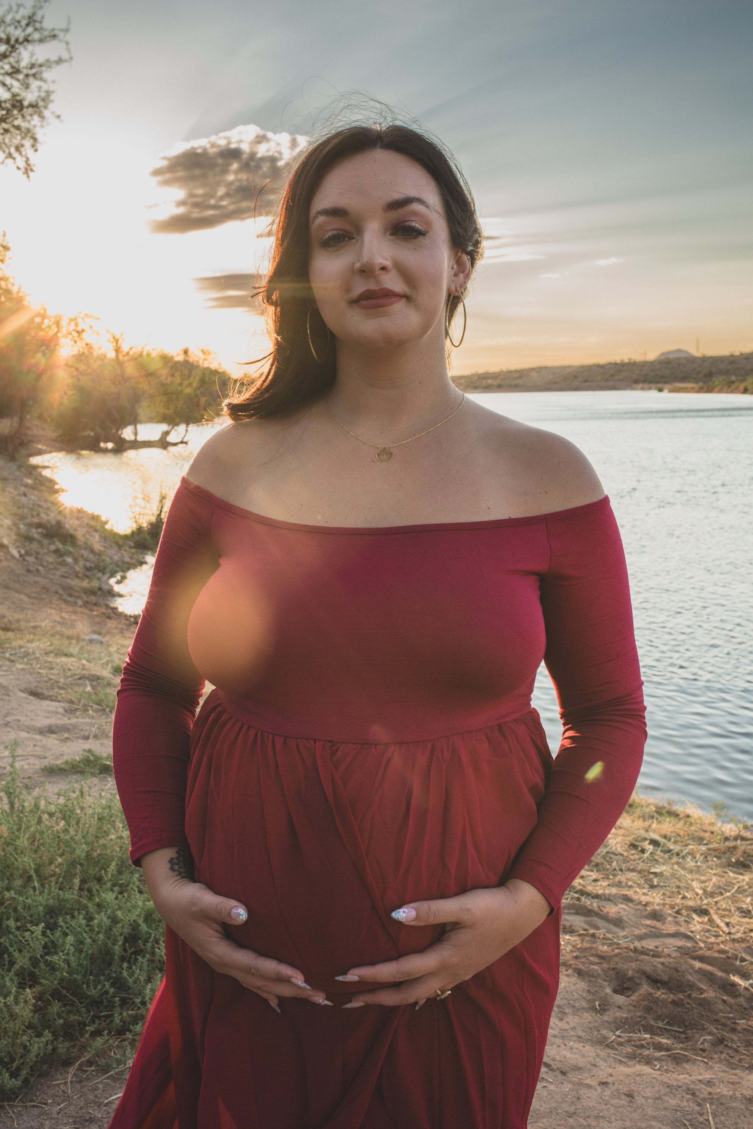 Mother-to-be's poses with her pregnant belly in a red dress for maternity photos in front of the Salt River and mountain by the best maternity photographer in Phoenix; Jennifer Lind Schutsky.