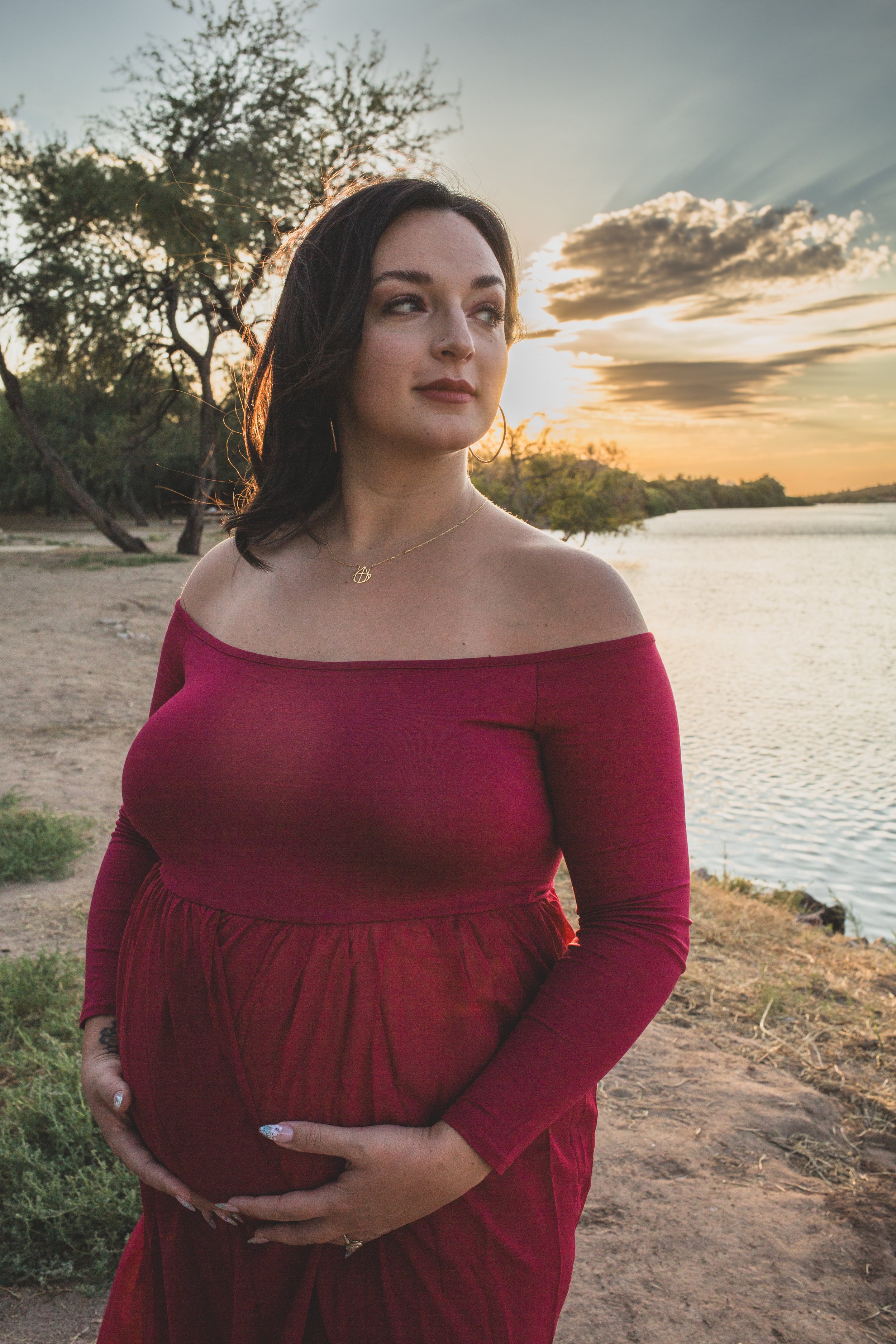 Mother-to-be's poses with her pregnant belly in a red dress at Sunset for maternity photos in front of the Salt River by maternity photographer in Phoenix; Jennifer Lind Schutsky.