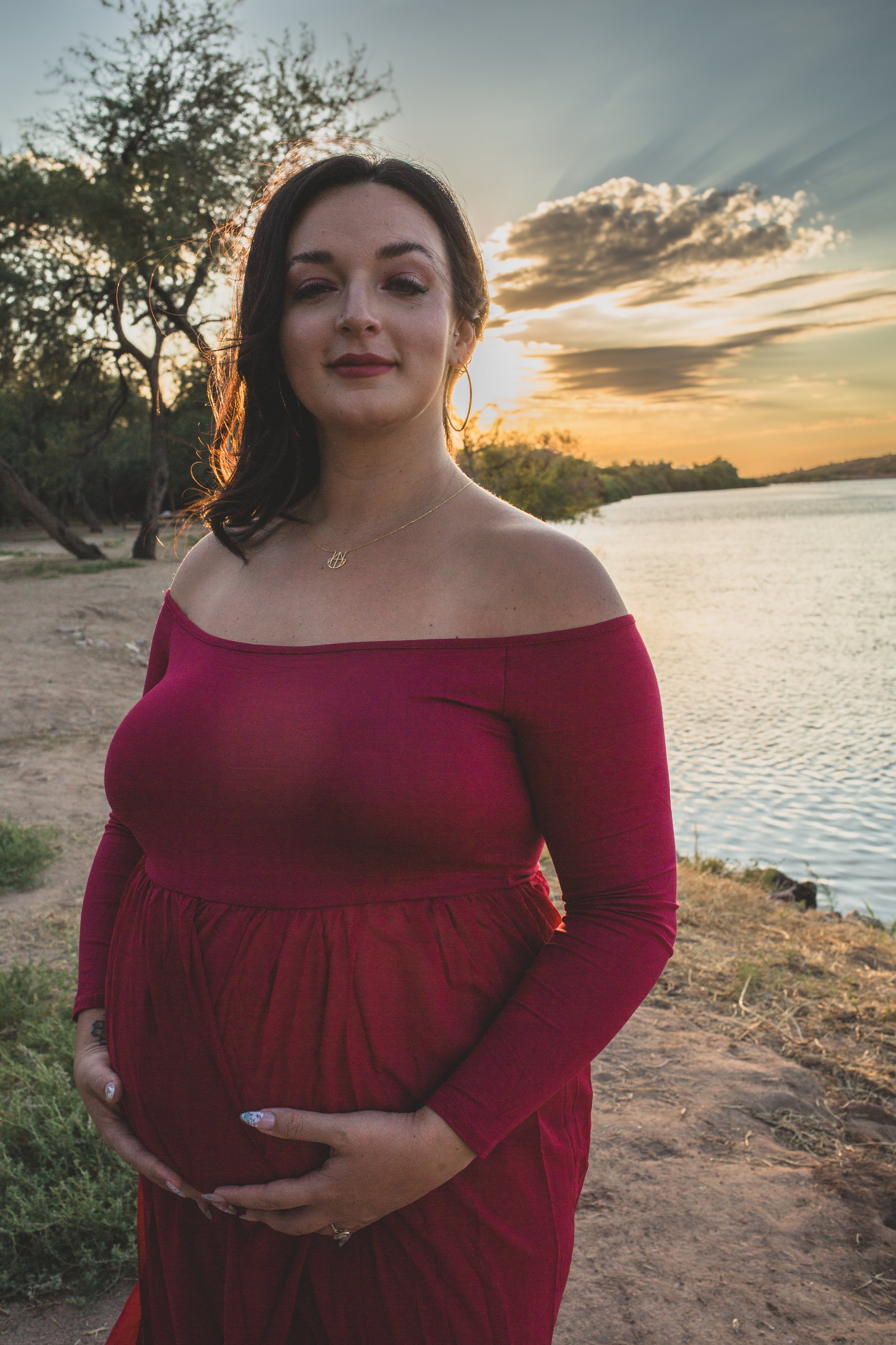 Mother-to-be's poses with her pregnant belly in a red dress at Sunset for maternity photos in front of the Salt River by Phoenix maternity photographer in Arizona; Jennifer Lind Schutsky.