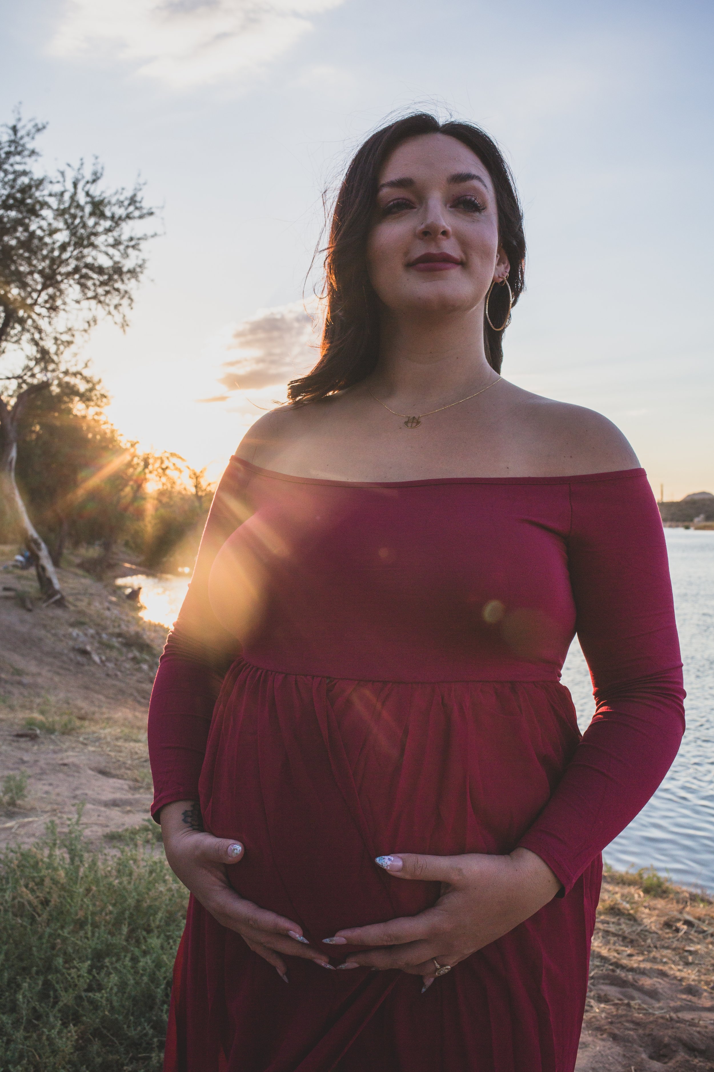 Mother-to-be's poses with her pregnant belly in a red dress at Sunset for maternity photos in front of the Salt River by Phoenix maternity photographer in Arizona; Jennifer Lind Schutsky.