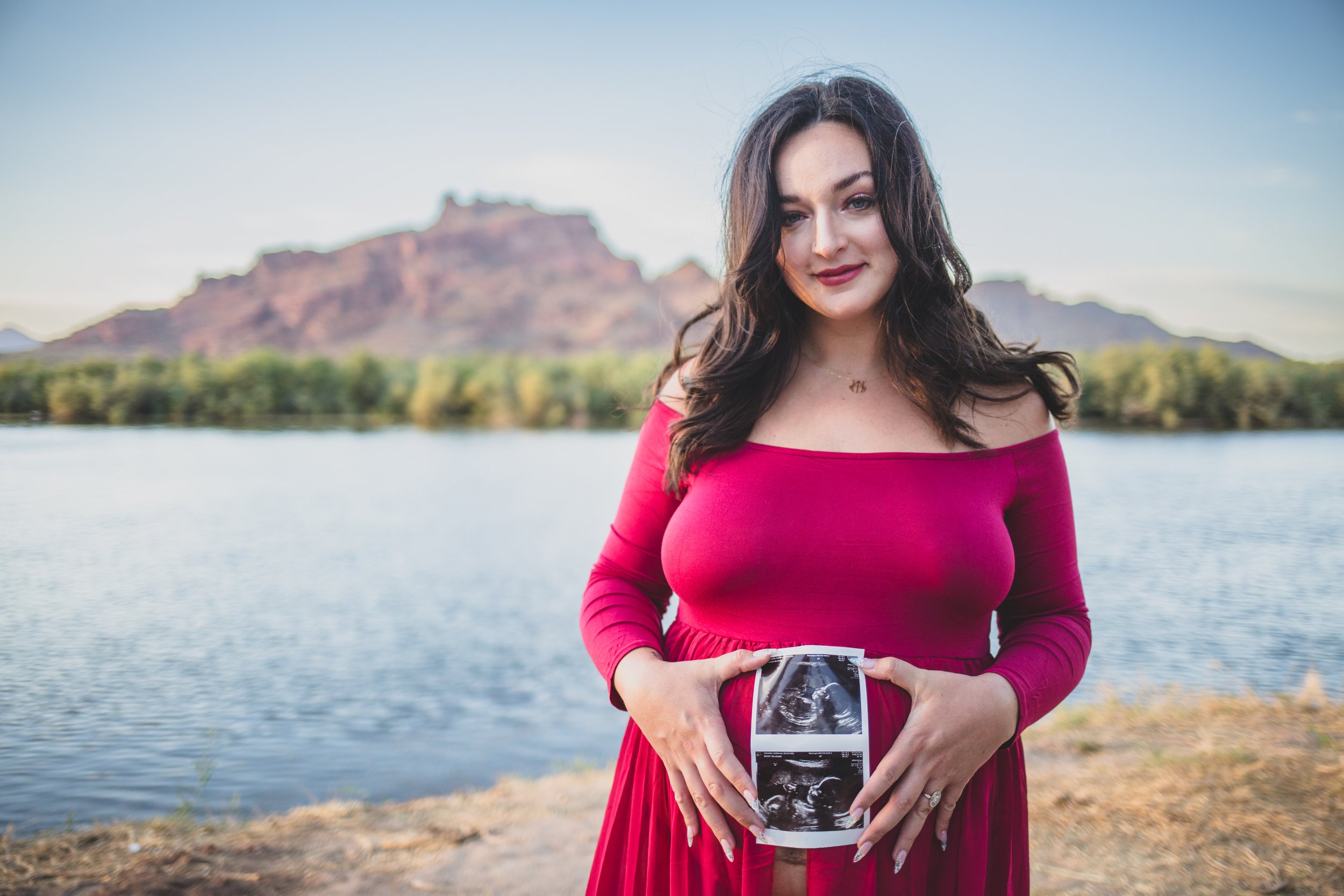 Expecting mother-to-be poses in a red dress with ultrasound pictures for maternity photos in front of the Salt River and mountain by timeless Arizona maternity photographer; Jennifer Lind Schutsky.