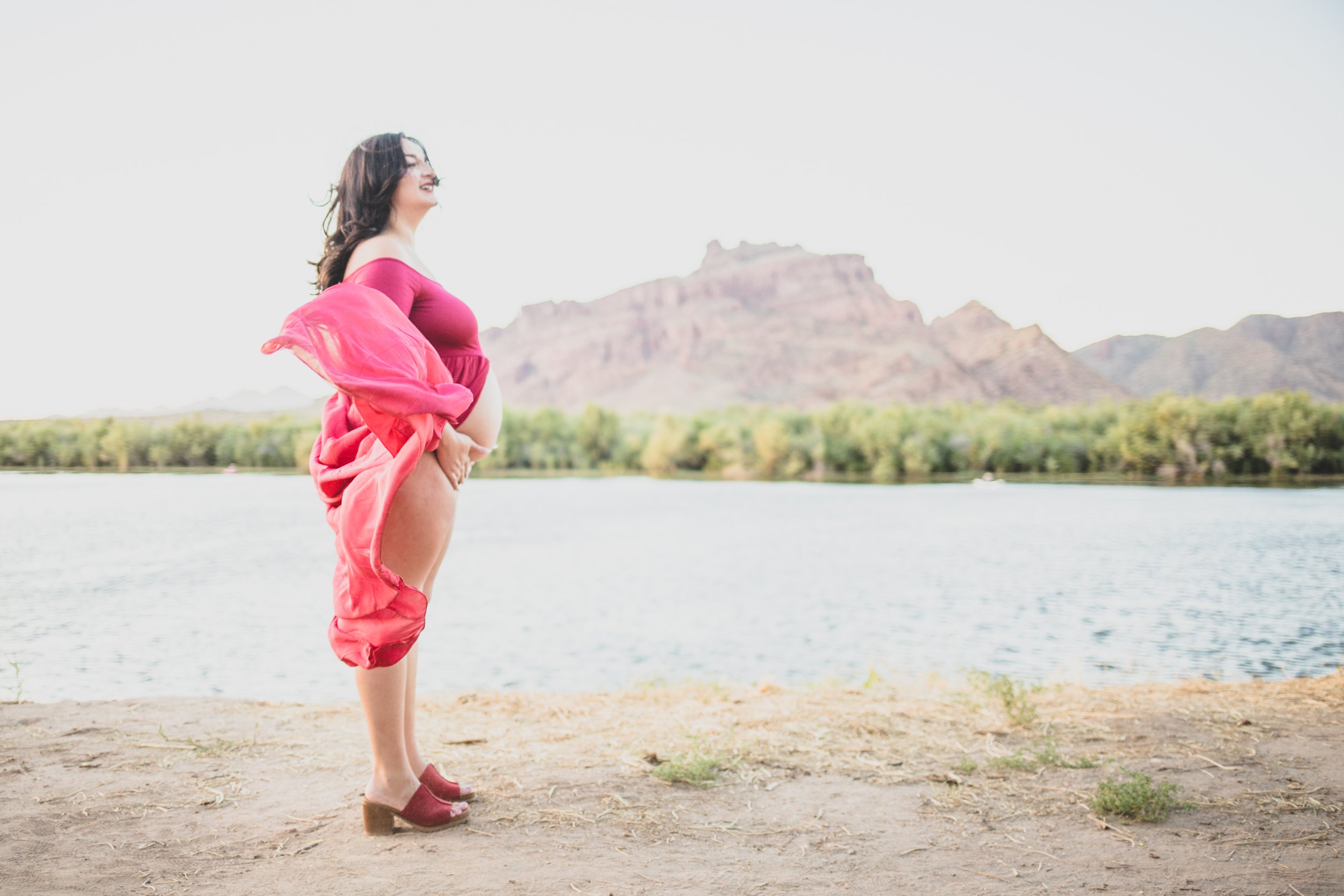 Woman poses in a red dress with for film style maternity photos in front of the Salt River and mountain by timeless Arizona maternity photographer; Jennifer Lind Schutsky.