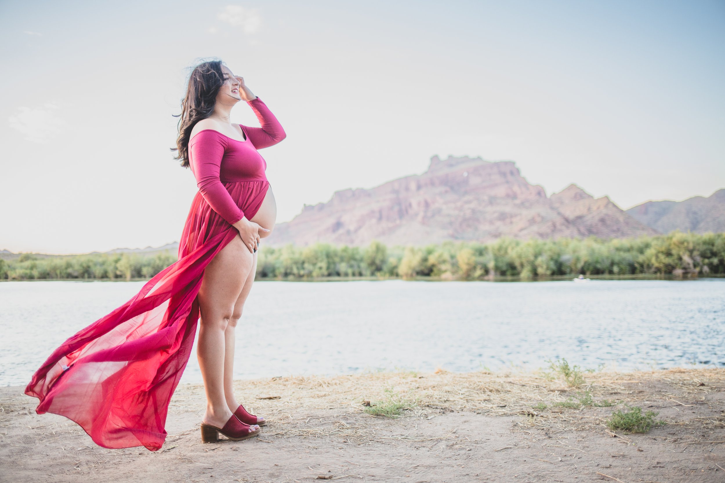 Woman poses in a red dress with for film style maternity photos in front of the Salt River and mountain by timeless Arizona maternity photographer; Jennifer Lind Schutsky.