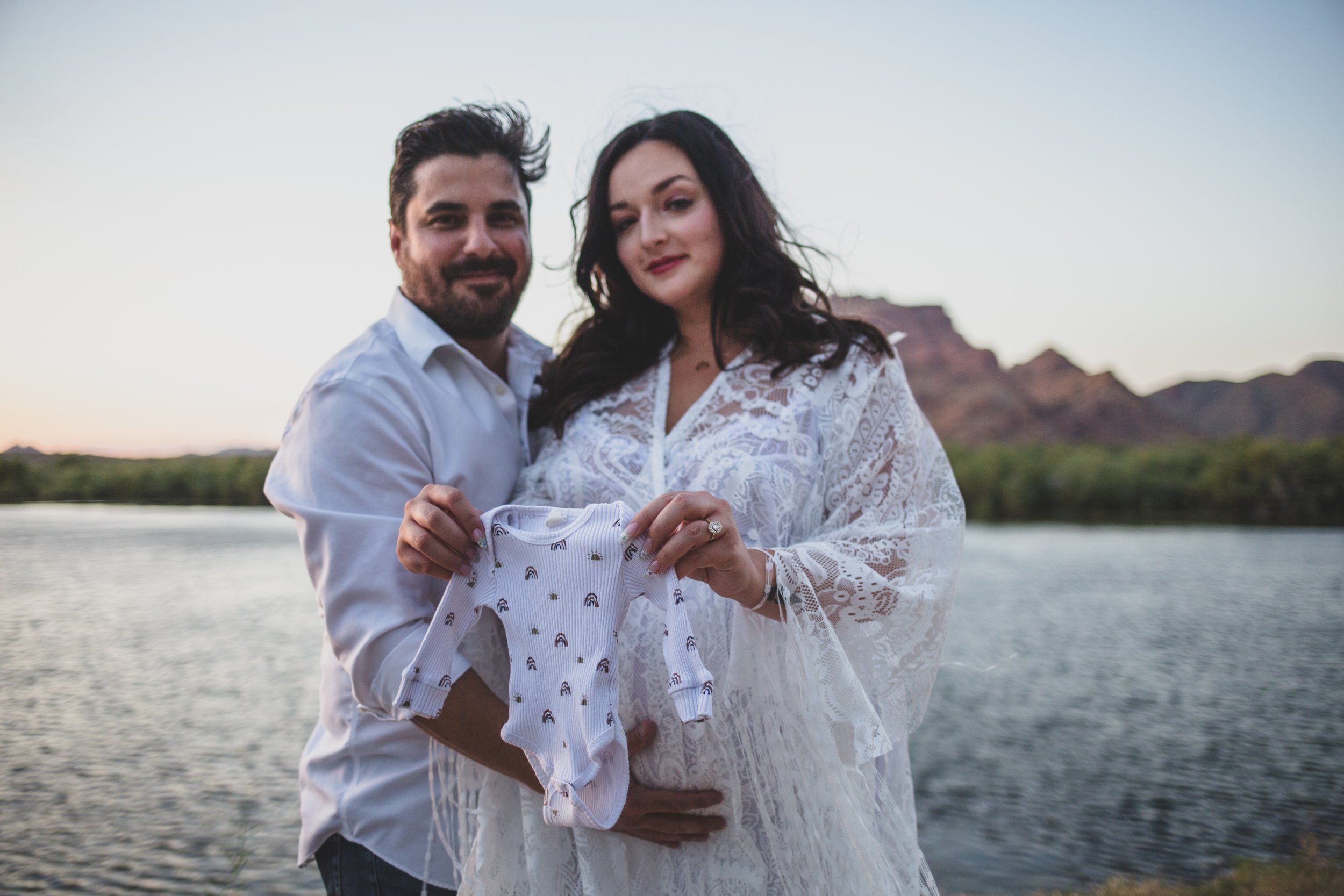 Romantic couple poses with newborn outfit for maternity photos near the water at the Salt River by timeless Phoenix maternity photographer; Jennifer Lind Schutsky.