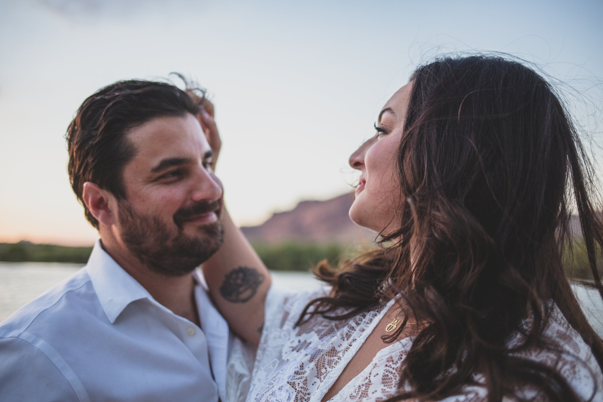Romantic couple poses with the wind in their hair for maternity photos near the water at the Salt River by timeless Phoenix maternity photographer; Jennifer Lind Schutsky.