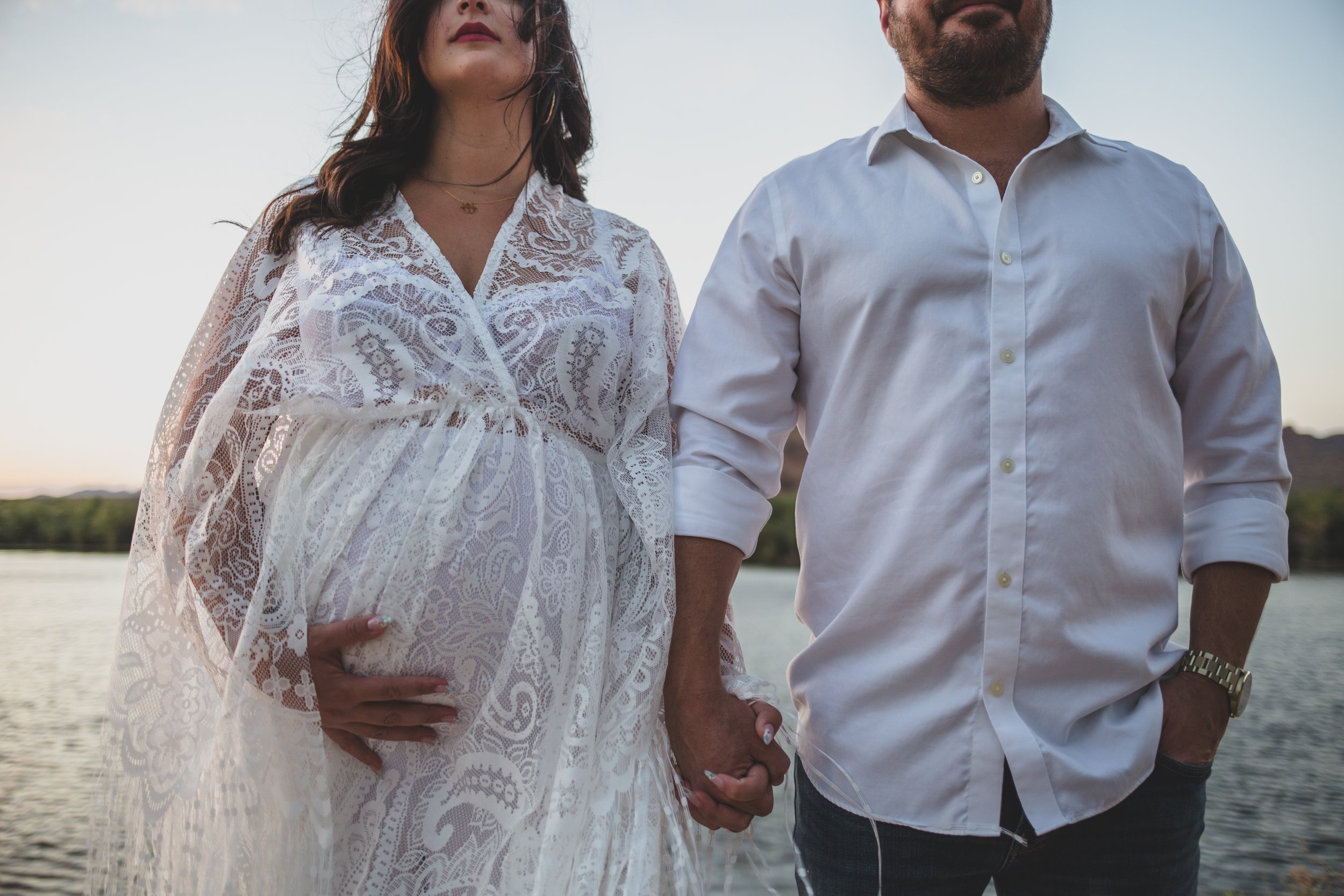 Romantic couple poses with the wind in their hair for maternity photos near the water at sunset the Salt River by timeless Phoenix maternity photographer; Jennifer Lind Schutsky.