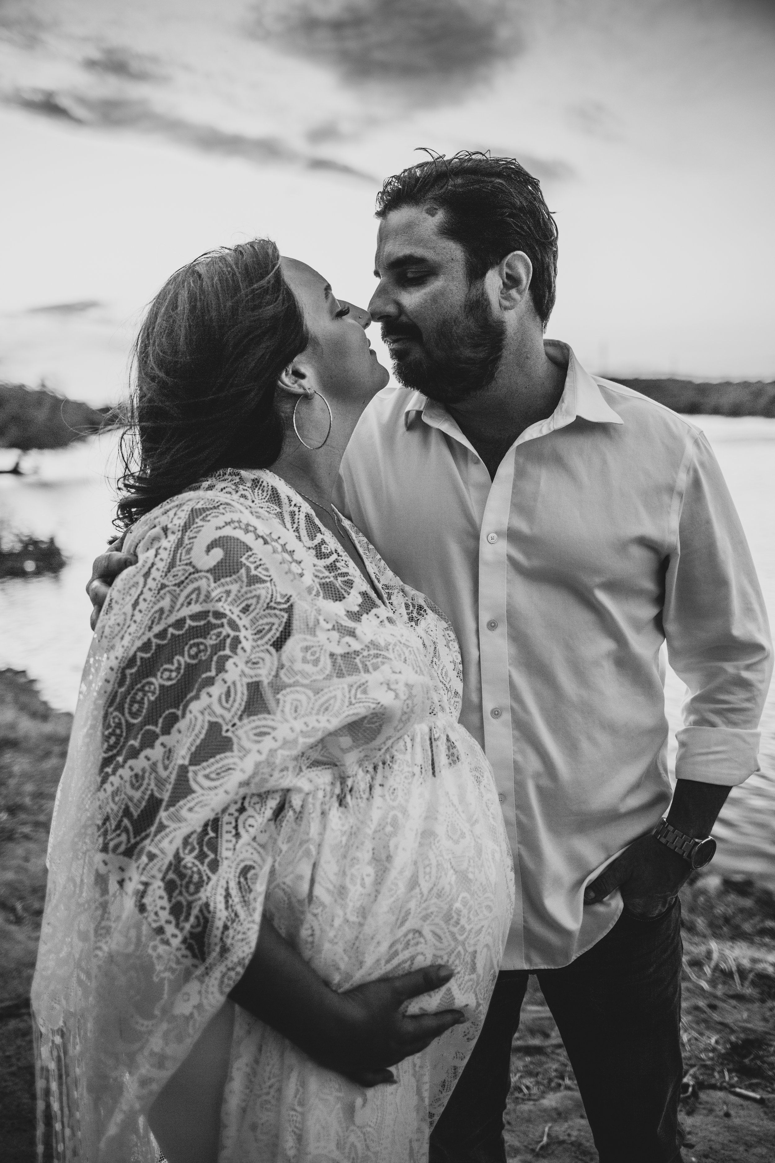 Romantic couple poses with the wind in their hair for maternity photos near the water at sunset the Salt River by timeless Phoenix maternity photographer; Jennifer Lind Schutsky.